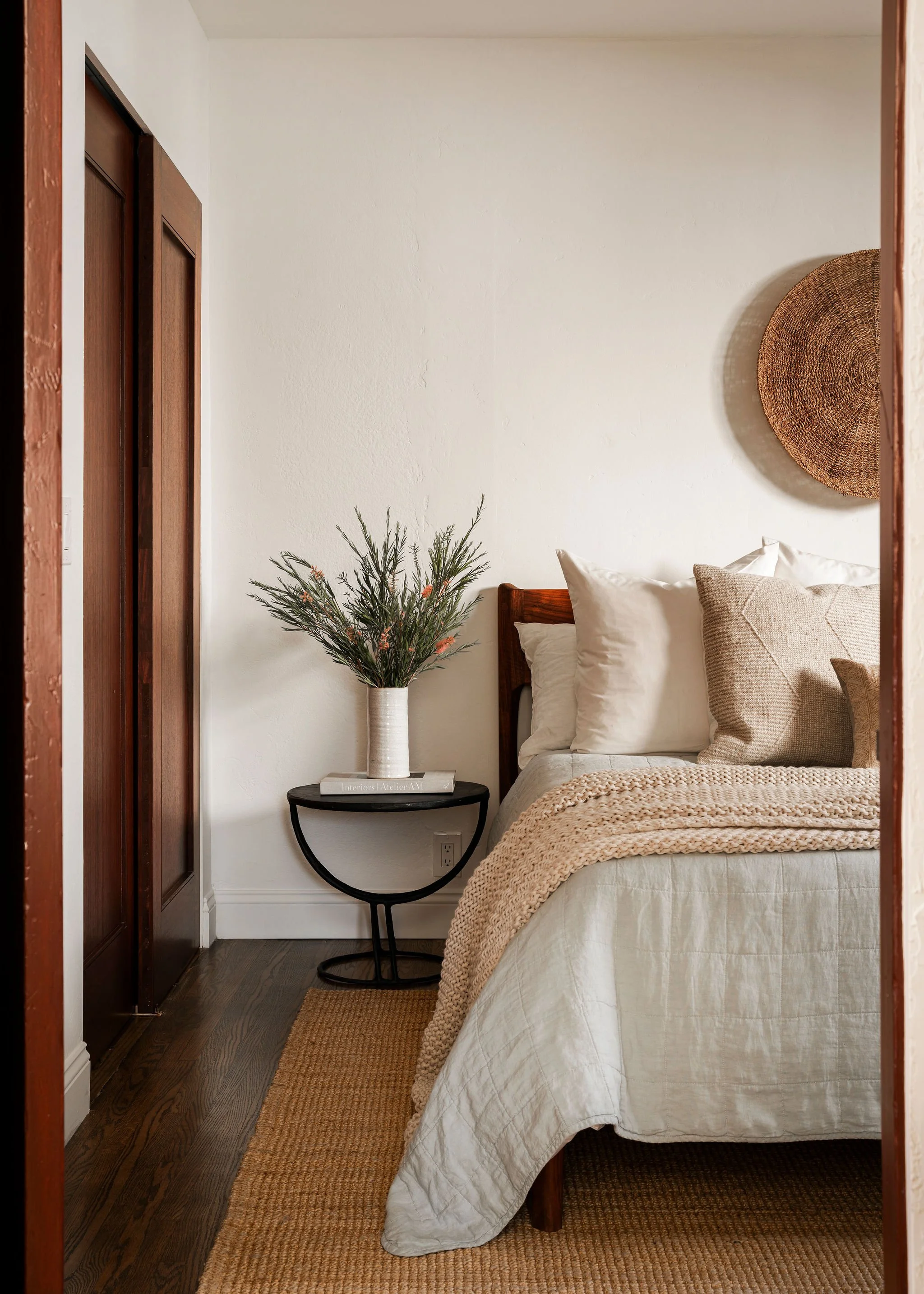 A cozy bedroom with a wooden bed frame, white bedding, beige throw blanket, and multiple pillows. A wicker wall decor hangs above the bed. To the left, there's a small black side table with a white vase holding greenery and small flowers, and a book 