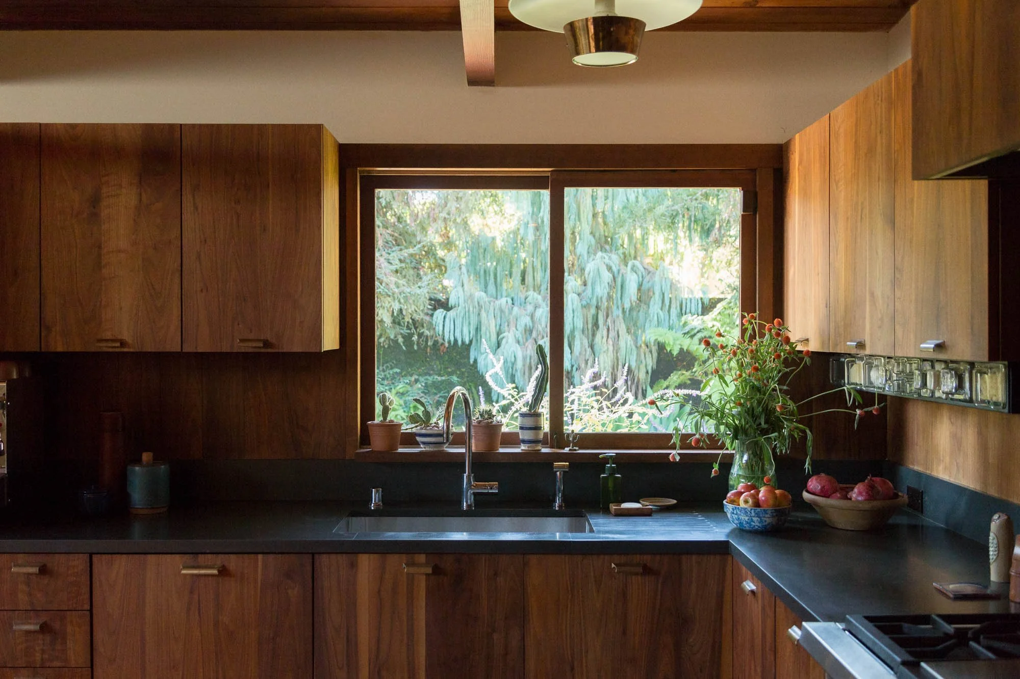 Kitchen with wooden cabinets, black countertop, window with a garden view, potted plants, a vase with flowers, bowls of apples, and a faucet.