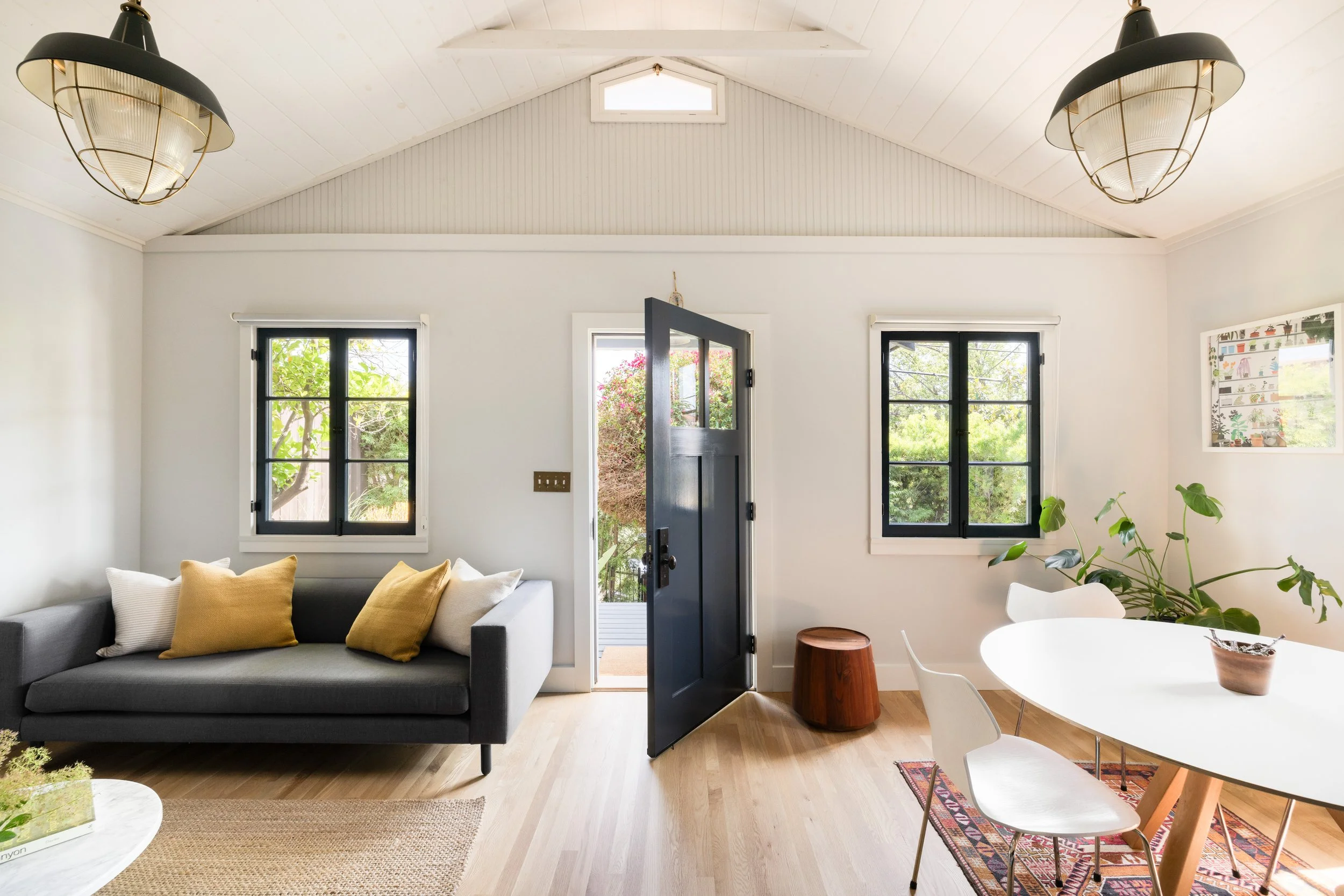 Living room with black door open to outdoor garden, two black-framed windows, gray sofa with white and beige pillows, dining table with white chairs, potted plant, and wooden stool, with light wooden flooring and white walls.