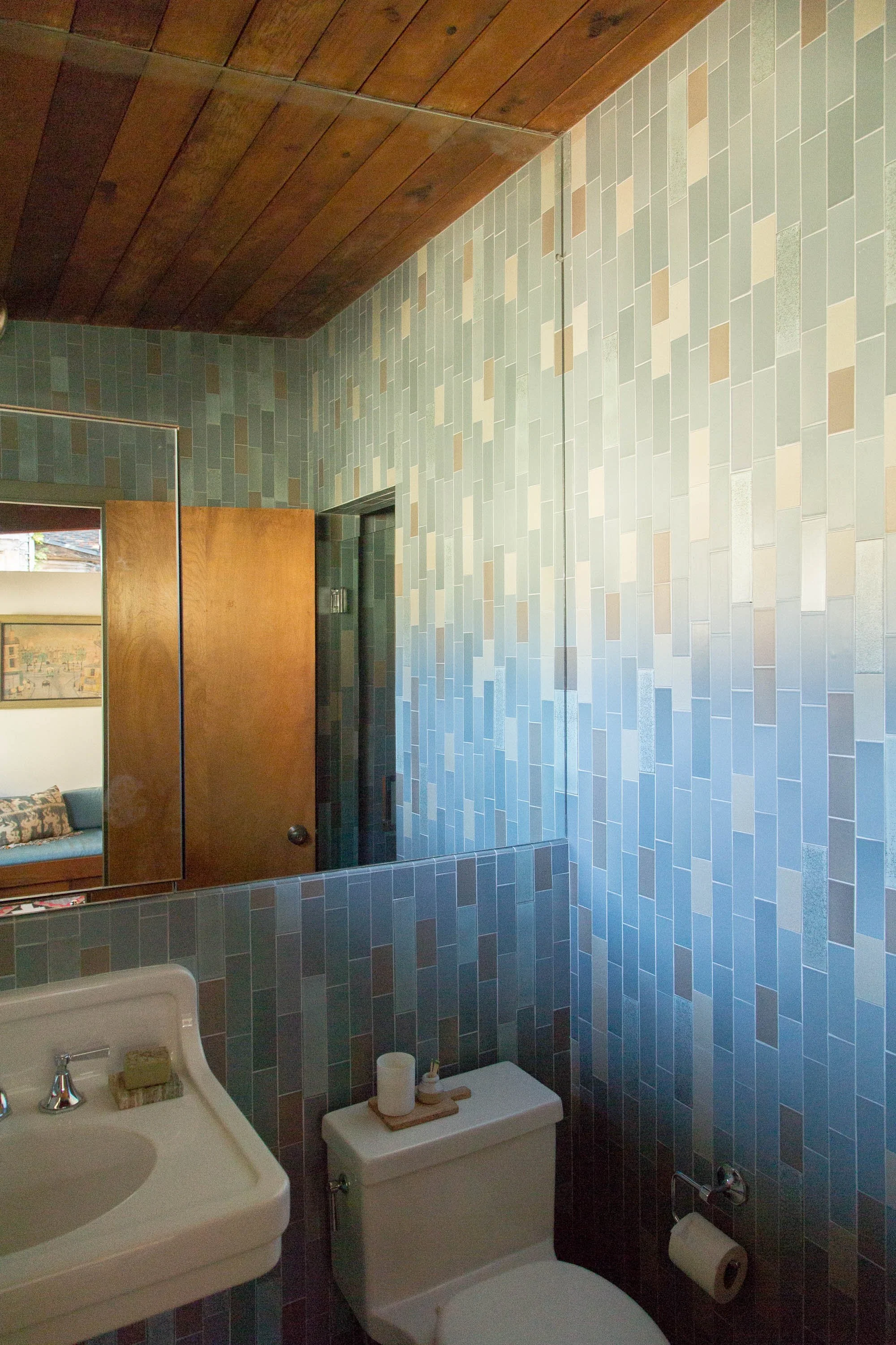Bathroom with a tiled wall in shades of blue, a toilet with a wooden tray on top holding a candle and soap, a sink with a bar of soap, a mirror, and a wooden ceiling.