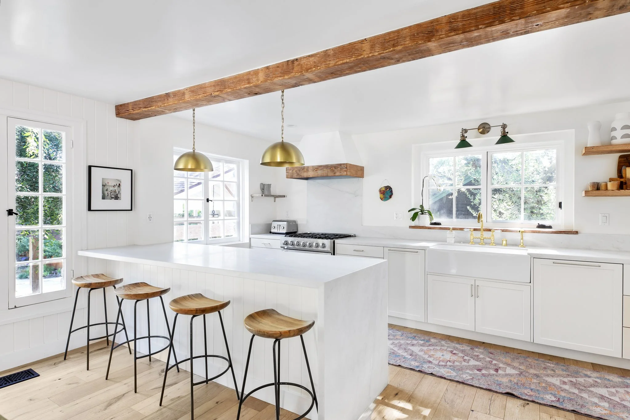 Bright white kitchen with wooden beams, gold pendant lights, and a large island with four wooden barstools. Features white cabinets, a window above the sink, and a door to the outside. Decor includes a framed picture and small wall art.