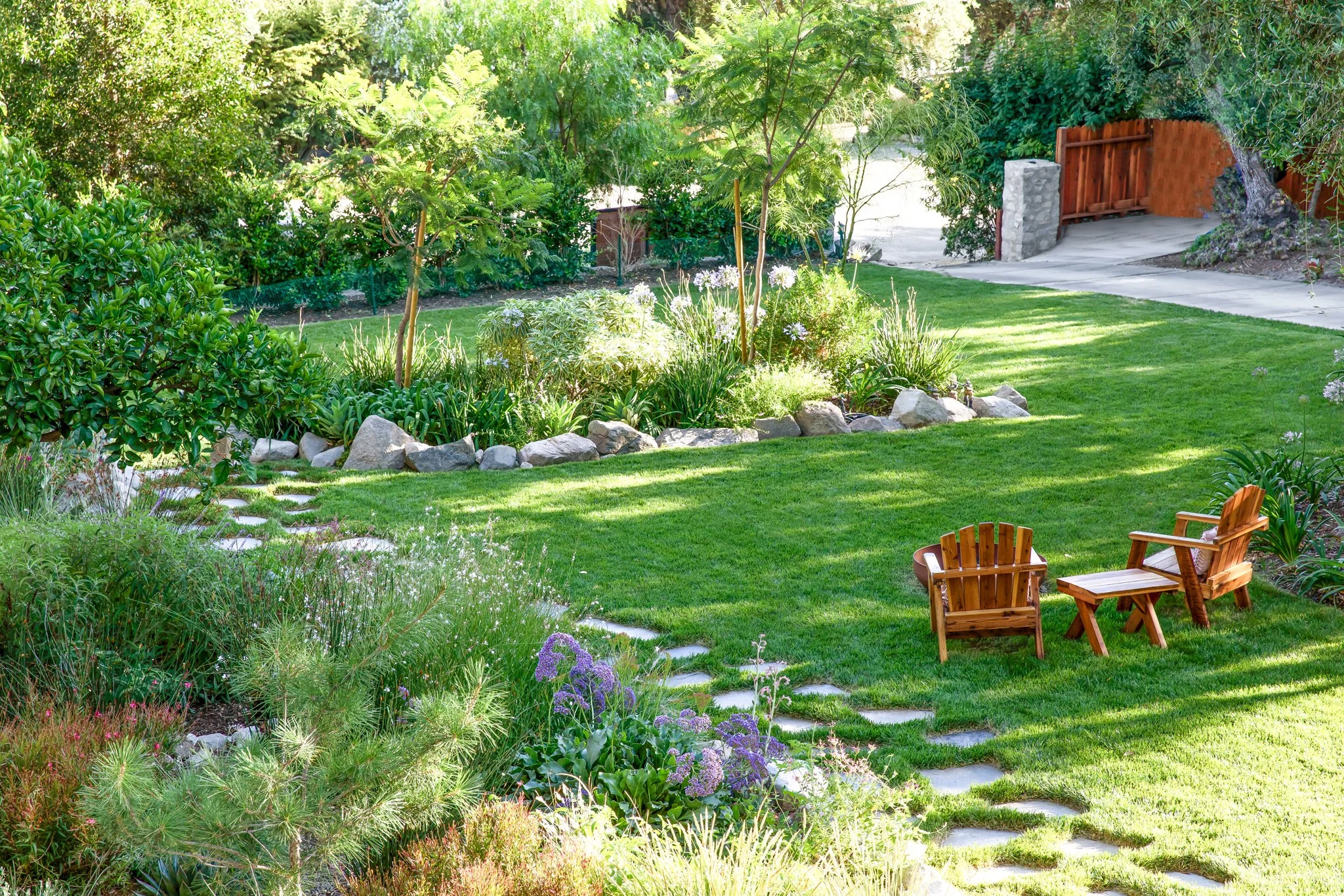 A lush green backyard with a small patio area containing two wooden Adirondack chairs, a garden bed with a variety of plants and flowers, a stone pathway, and a wooden gate in the background.