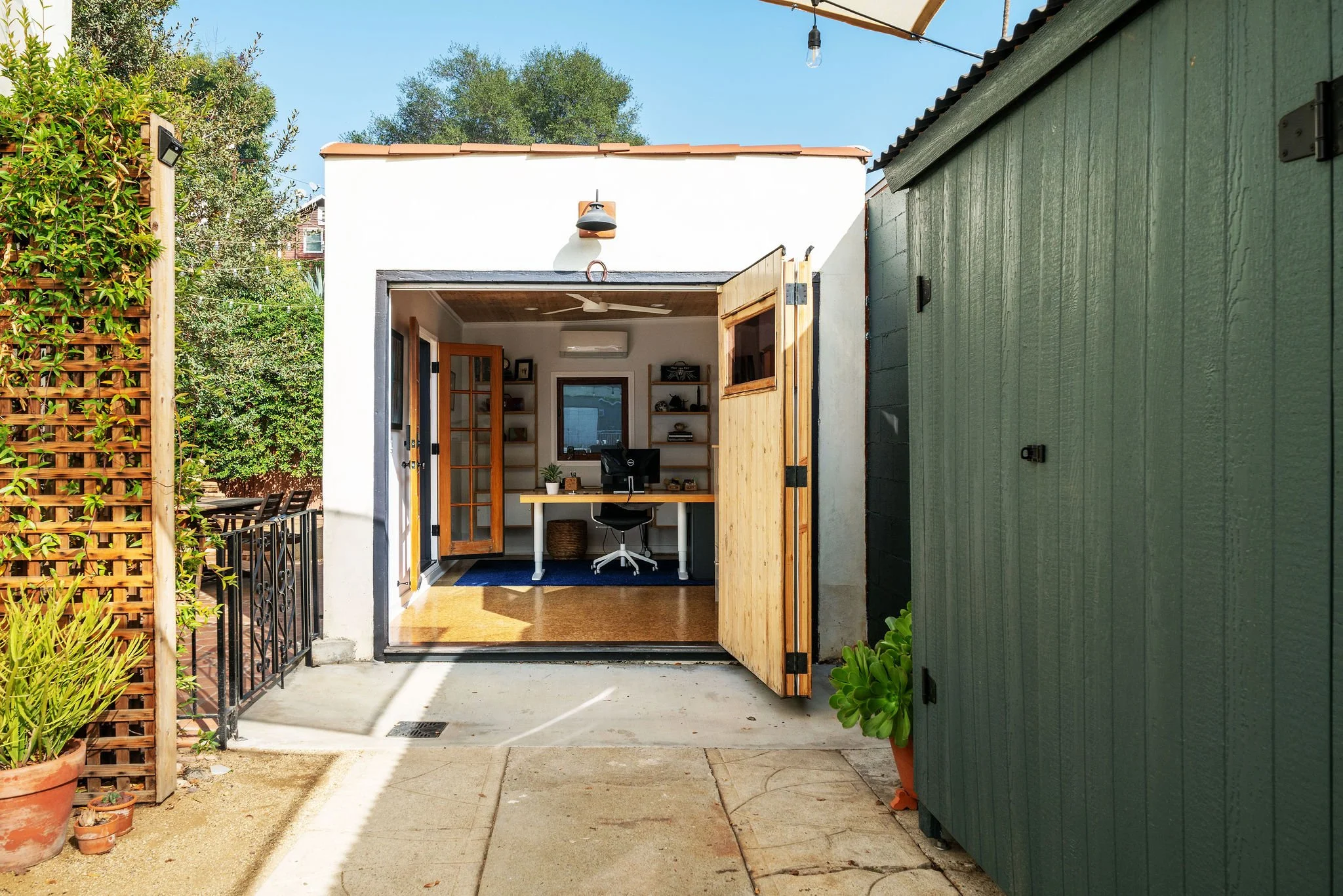 Open door leading to a small home office with a desk, chair, bookshelves, and window, set in a backyard with plants and a green shed.