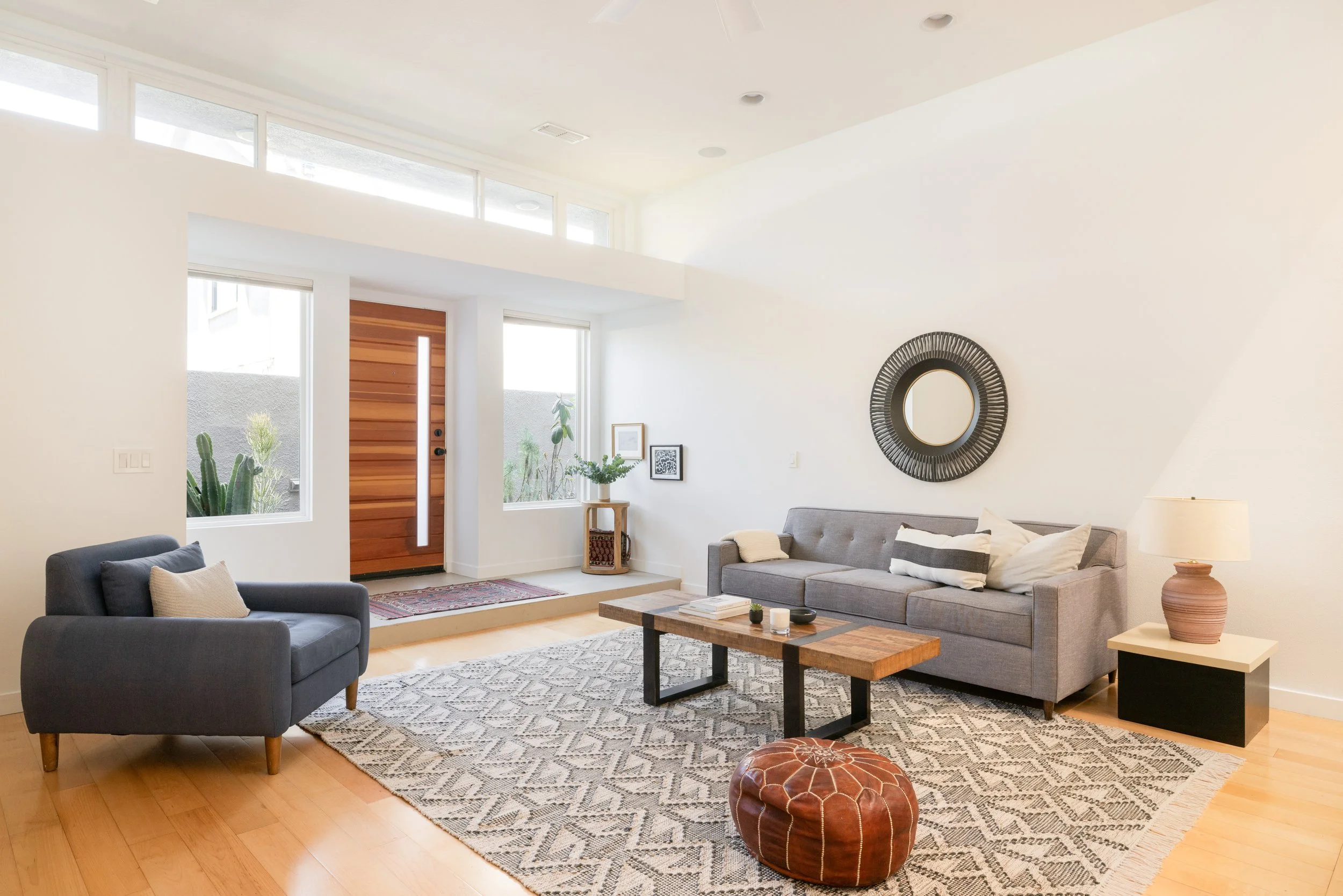 Living room with gray sofas, a wooden coffee table, patterned area rug, leather pouf, side table with lamp, decorative mirror, and front door with window.