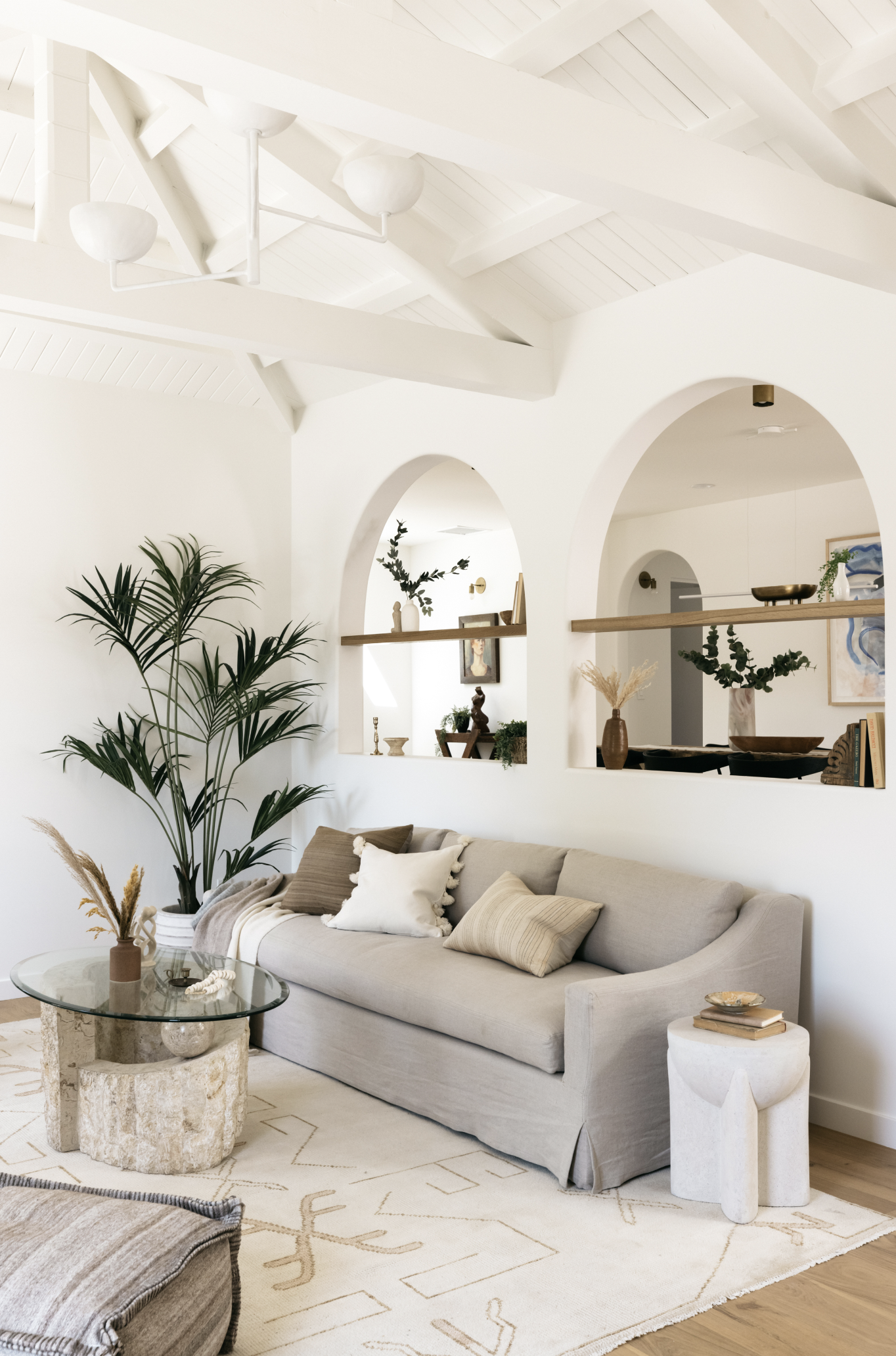 A minimalist living room with a beige sofa, a glass coffee table made of a stone base, and a large potted palm. The room has white walls, arched openings, and a vaulted white ceiling with exposed beams. Decor includes vases, books, and art pieces, cr