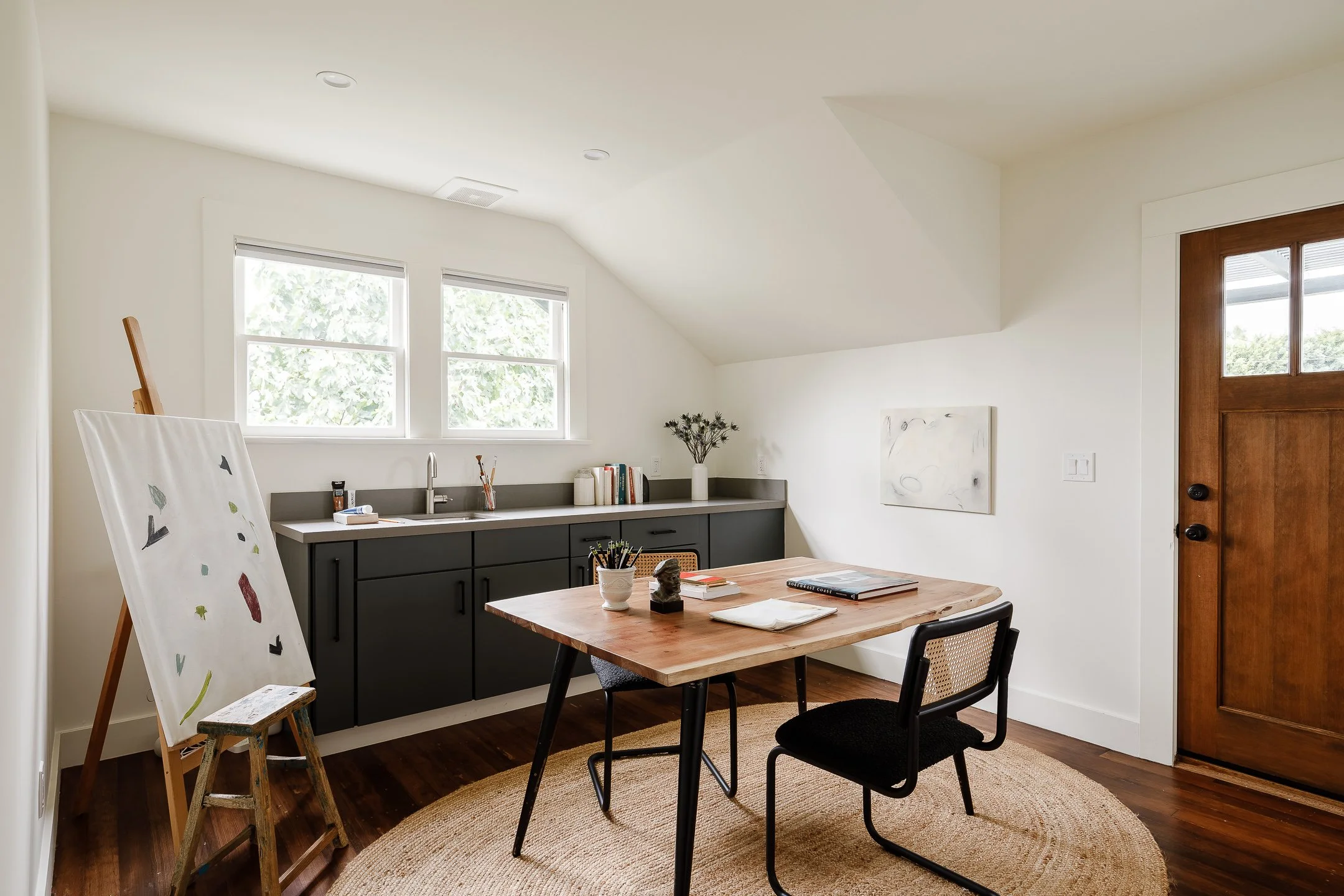 A modern home art studio with a wooden table, two black chairs, a black cabinet with a sink, large windows, an easel with a partially painted canvas, some books, painting supplies, a potted plant, and wall art, all in a well-lit room with hardwood fl
