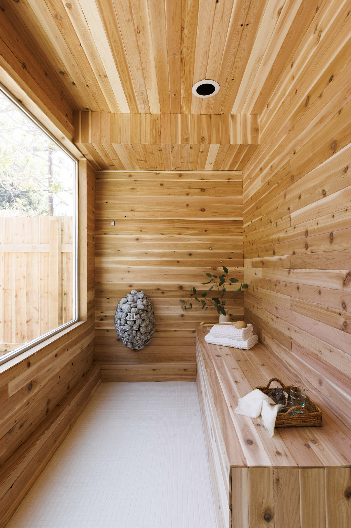 A wooden sauna with a wooden bench on the right, a plant, and a wicker basket with towels and bath accessories. A large window on the left lets in natural light.