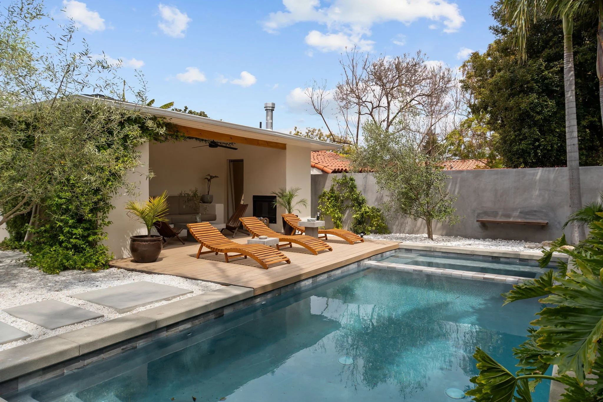 A backyard swimming pool with a wooden deck, lounge chairs, potted plants, a seating area, and a privacy wall. There are trees and a partly cloudy sky in the background.