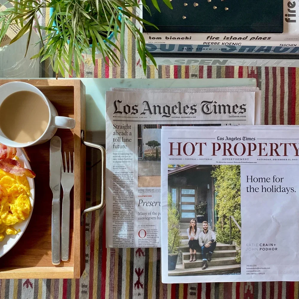 A breakfast scene with a plate of scrambled eggs and bacon, a cup of coffee, newspapers, and a pile of books on a table with a colorful striped tablecloth. A green plant is in the background.