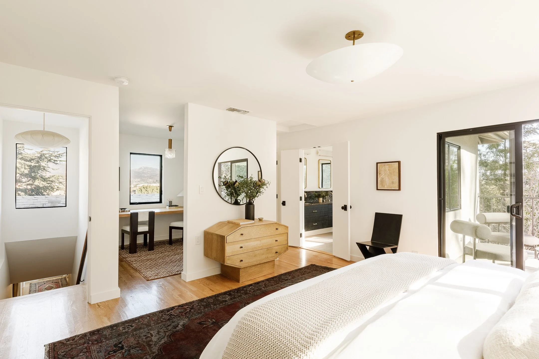 Bright bedroom with white walls, wooden floor, sliding glass door overlooking nature, bed with white bedding, black chair, decorative dresser, large mirror, and artwork.