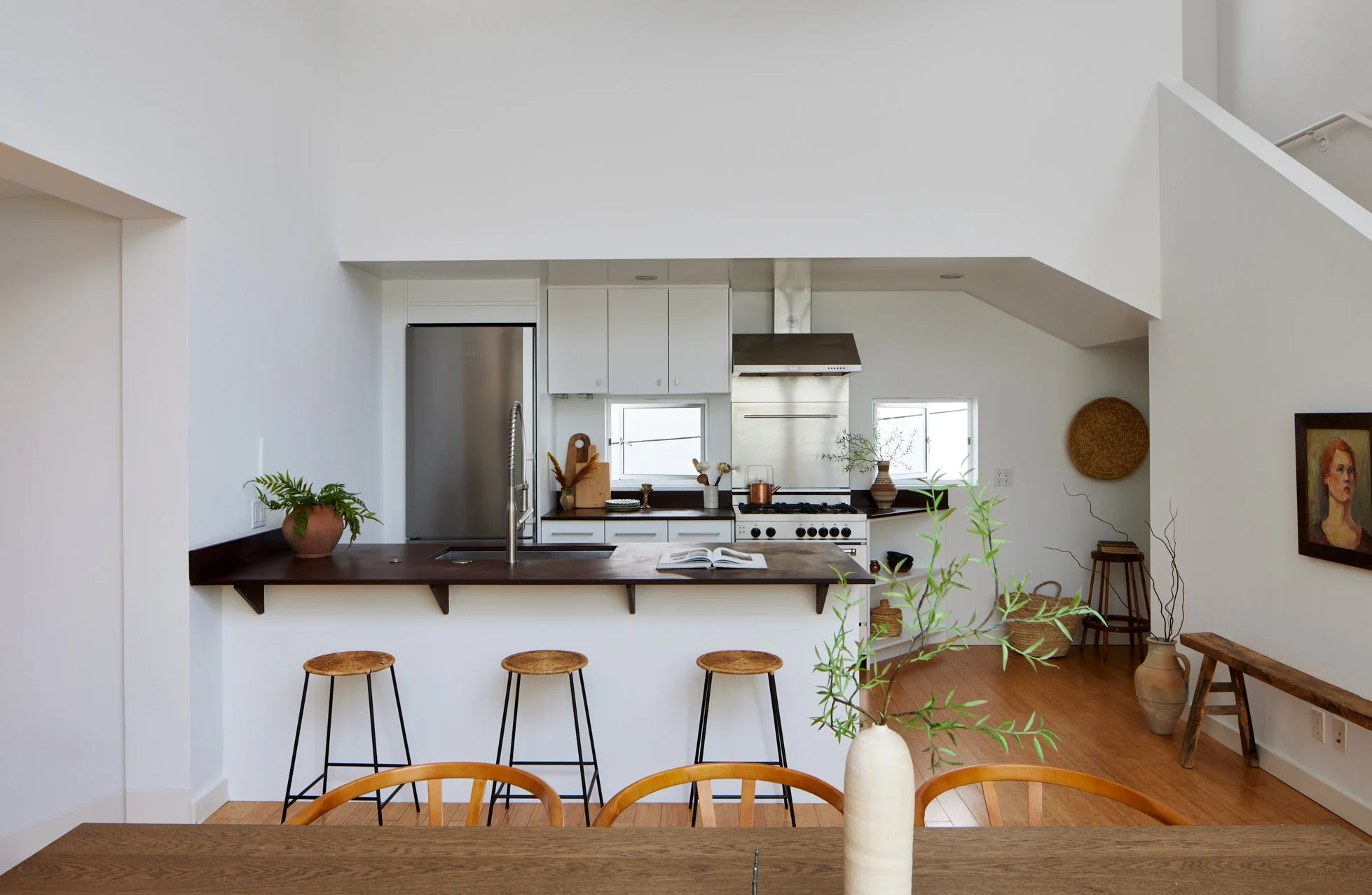 Modern kitchen with white walls, dark wood countertops, stainless steel refrigerator, and a stove. Small windows and decorative plants, with a wooden dining table and chairs in the foreground.