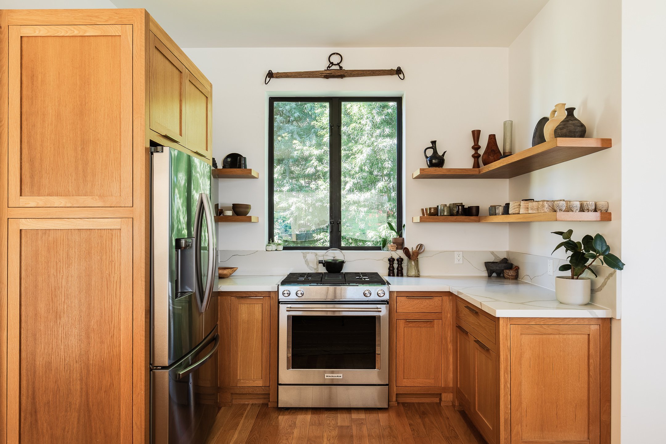 Kitchen with wooden cabinets, a stainless steel refrigerator, a stove under a window, and wooden floating shelves with decorative items.