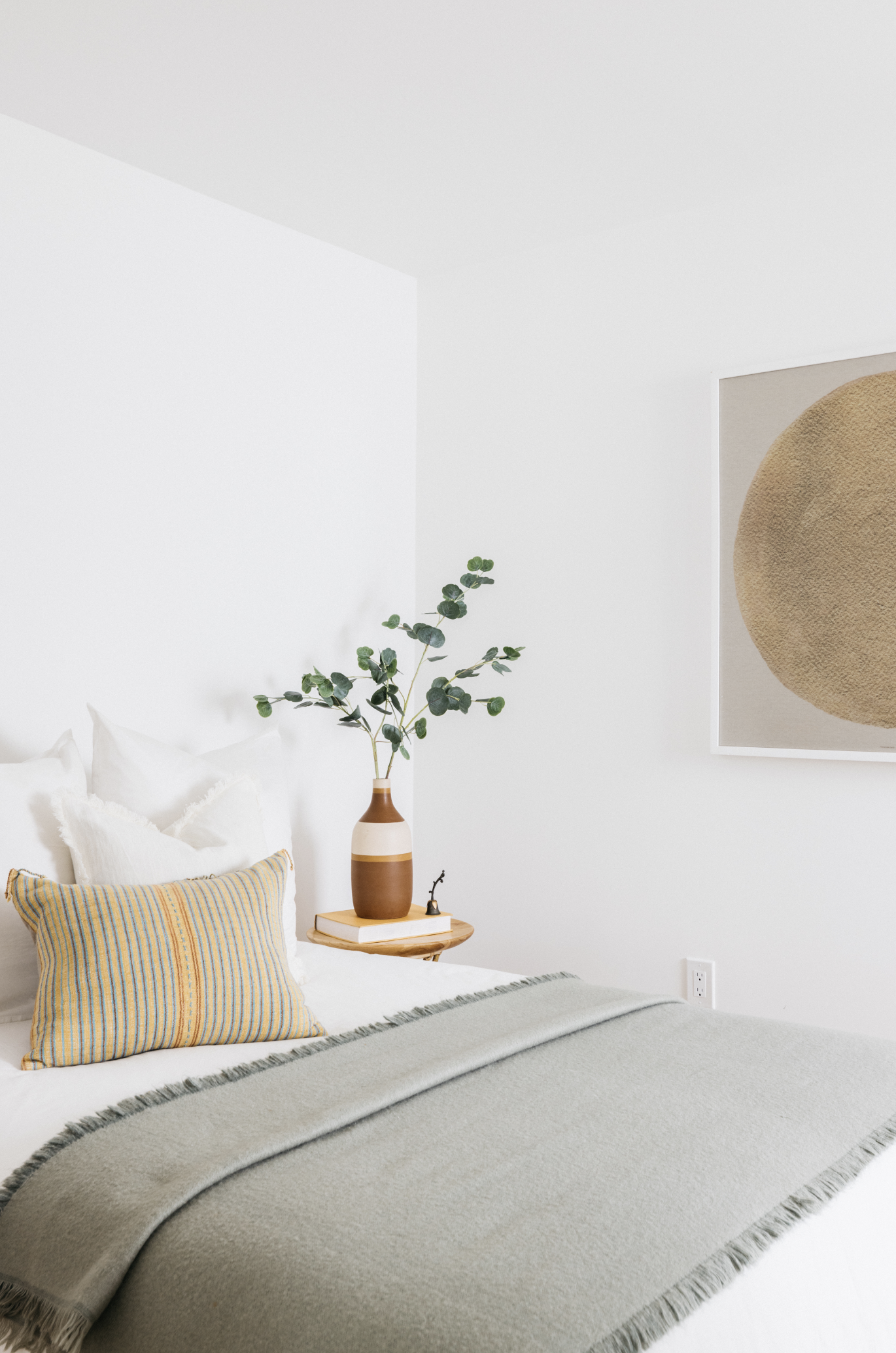 A neatly made bed with white pillows, a striped yellow and gray pillow, and a green throw blanket. A wooden nightstand with a beige and white vase holding green foliage, a small black figurine, and a stack of books. A large round artwork on the white