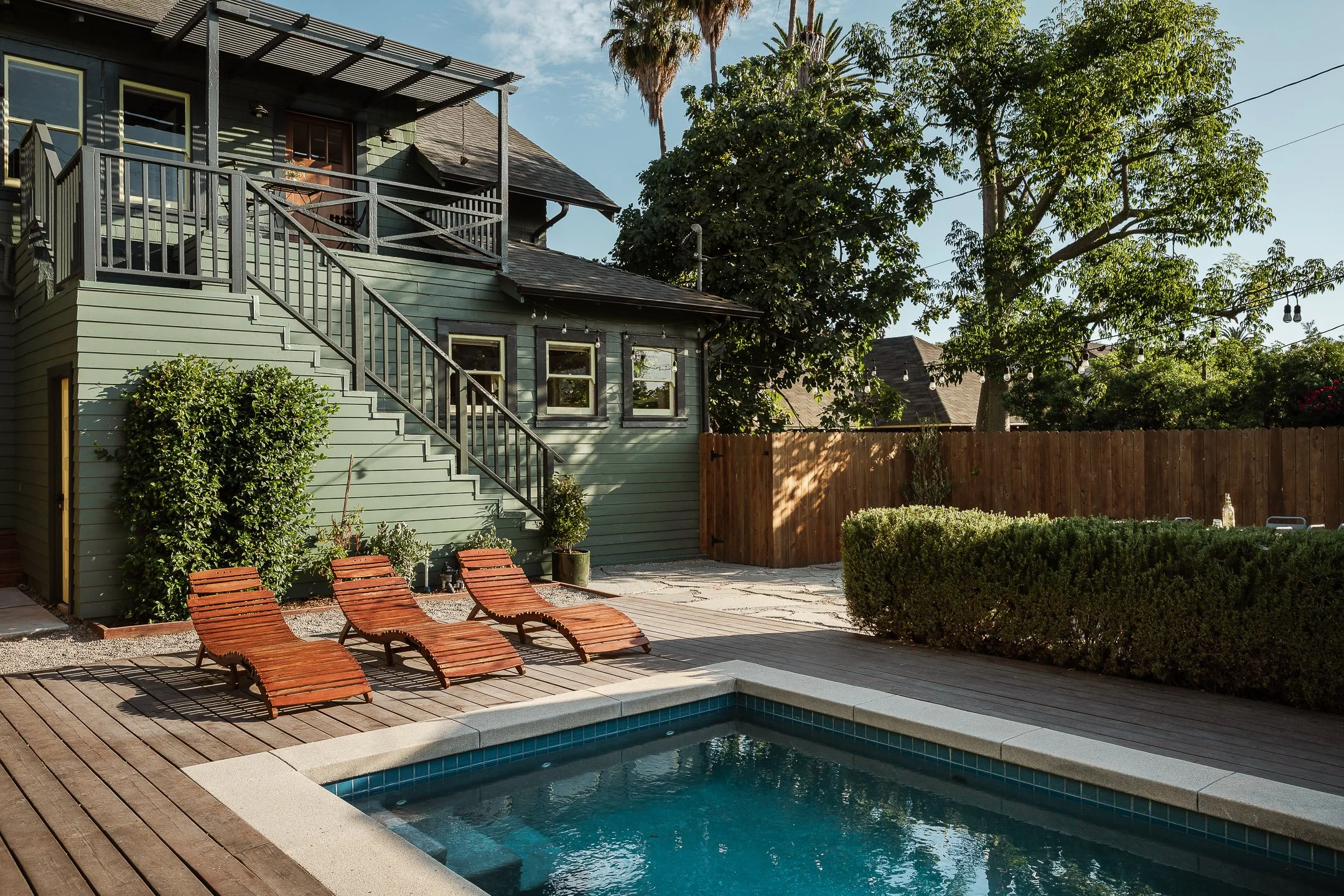Backyard with a small swimming pool, three wooden lounge chairs, a green house with stairs, trees, and a wooden fence.