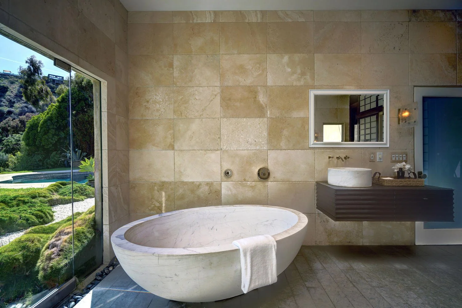 Modern bathroom with a oval white bathtub near a large glass window showing an outdoor landscape with greenery, rocks, and a pool.
