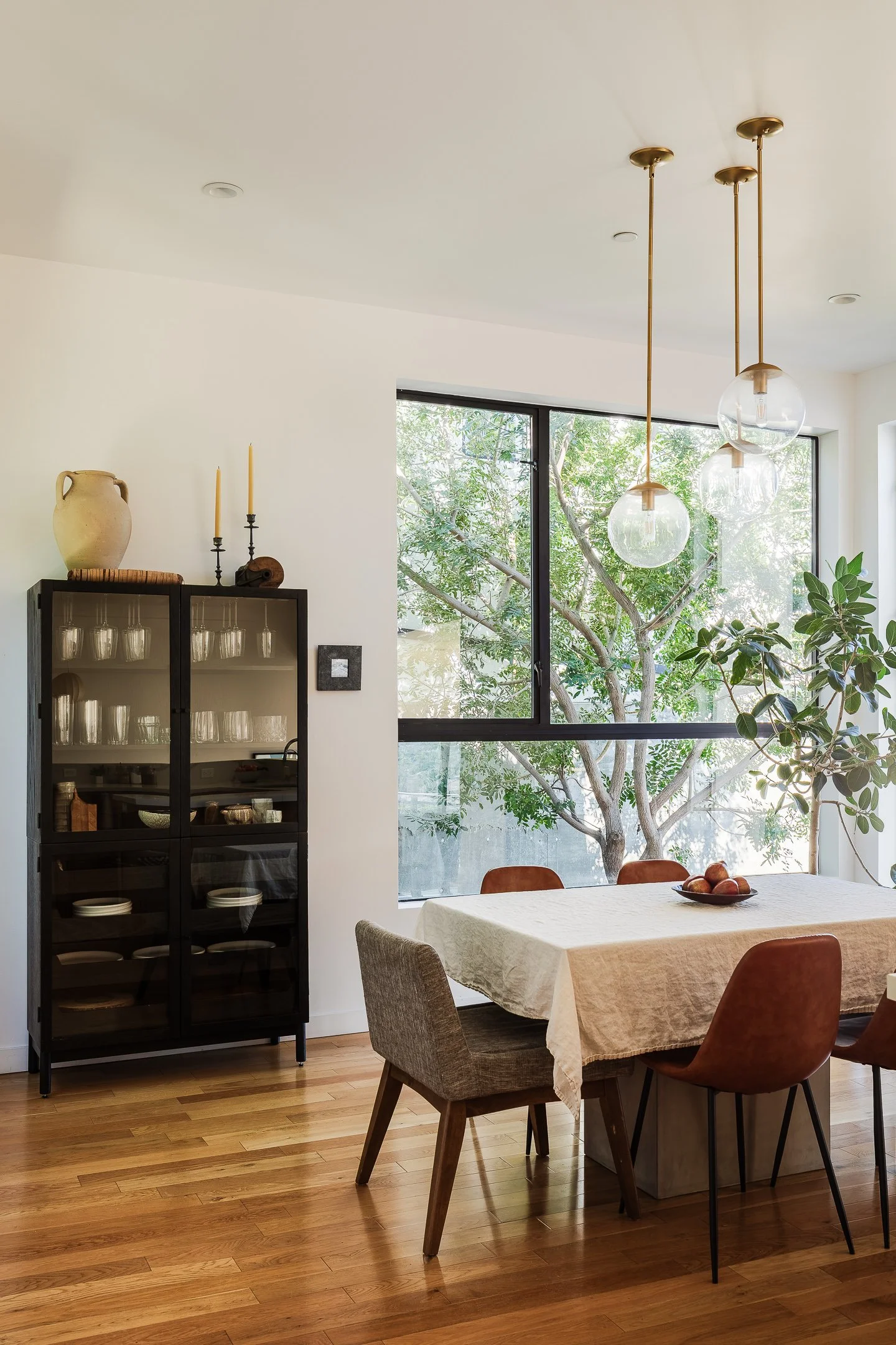 Modern dining room with large window, wooden floor, glass pendant lights, wooden dining table with beige tablecloth, and diverse chairs, with a view of green trees outside.