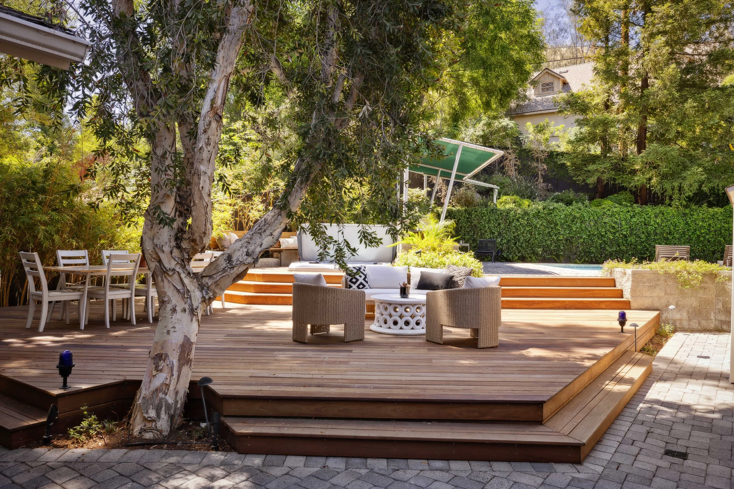 A spacious backyard patio with wooden decking, outdoor furniture including a round wicker chair set and a white sofa, surrounded by lush trees and greenery, with stairs leading up to a higher level, and a canopy providing shade over part of the space