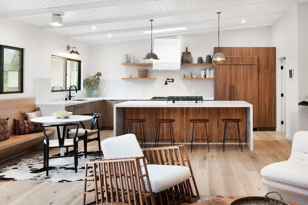 Open-concept kitchen and living room with white walls, wooden accents, and modern decor, including a kitchen island with three stools, a round dining table with chairs, and cozy seating area with chairs and a rug.