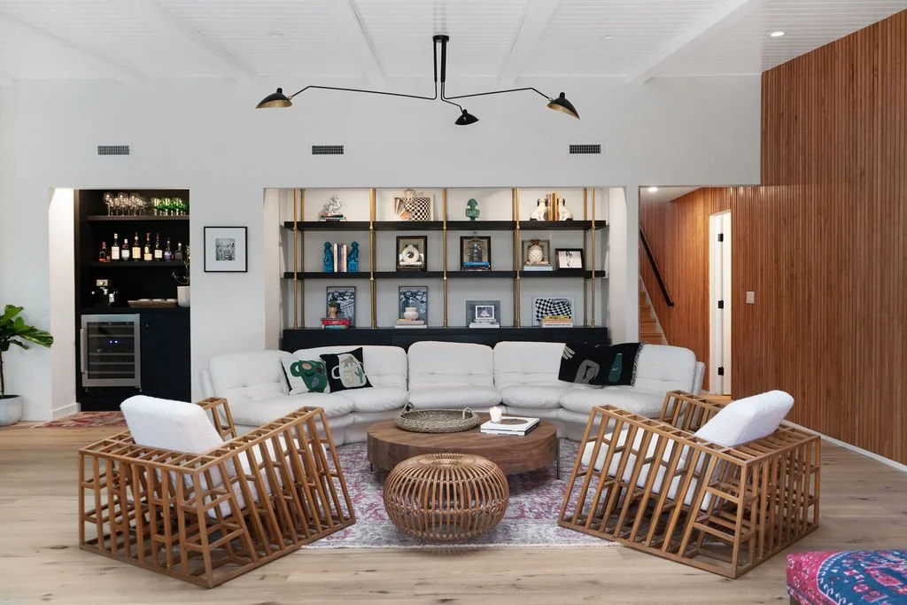 Modern living room with white sectional sofa, wooden armchairs, a round wood coffee table, decorative shelves with art and decor, and a built-in bar with liquor bottles in the corner.
