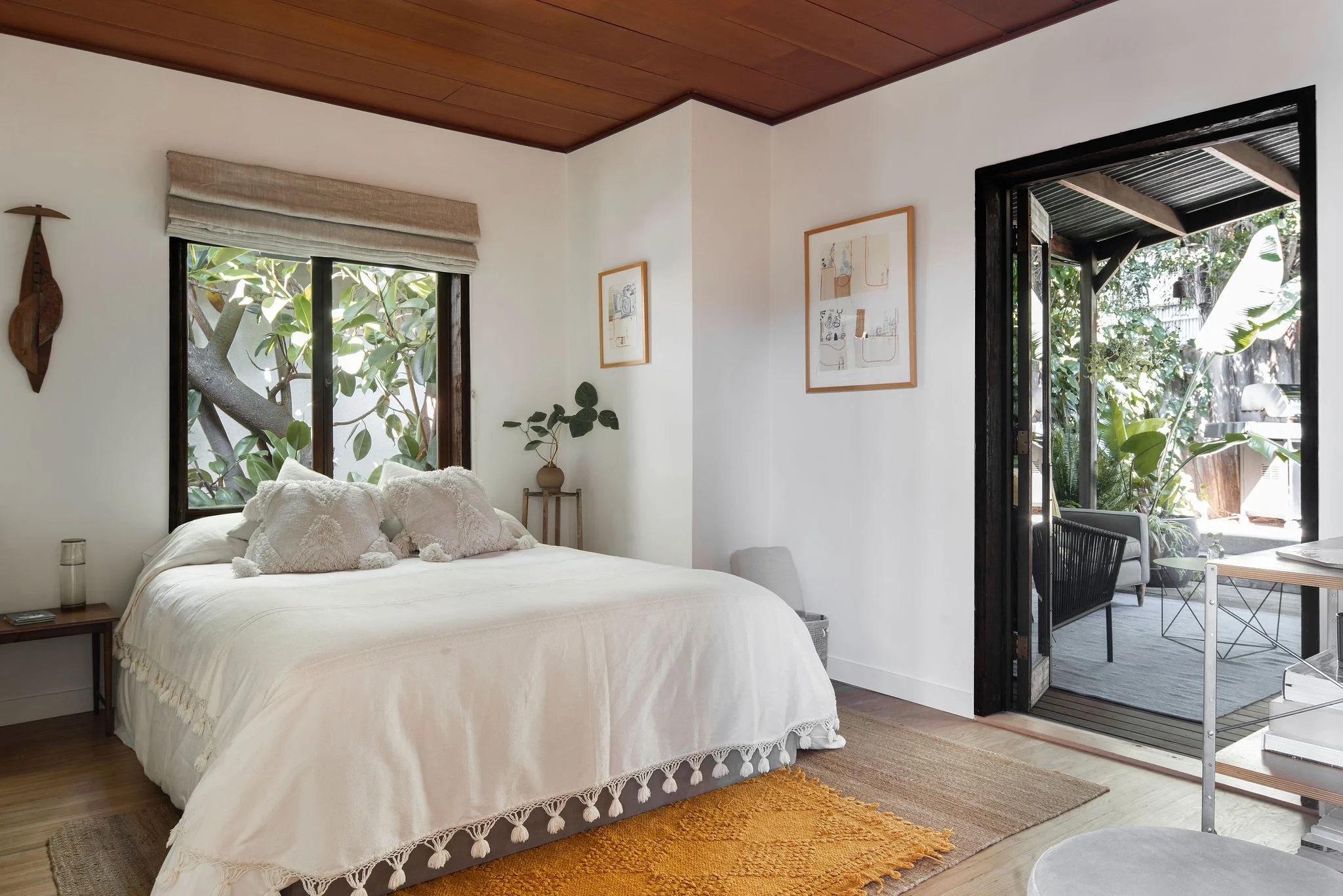 A bedroom with a white bed, decorative pillows, a side table with a glass, a potted plant, framed artwork on the wall, a window with greenery outside, and a sliding door opening to a patio with outdoor furniture and lush plants.