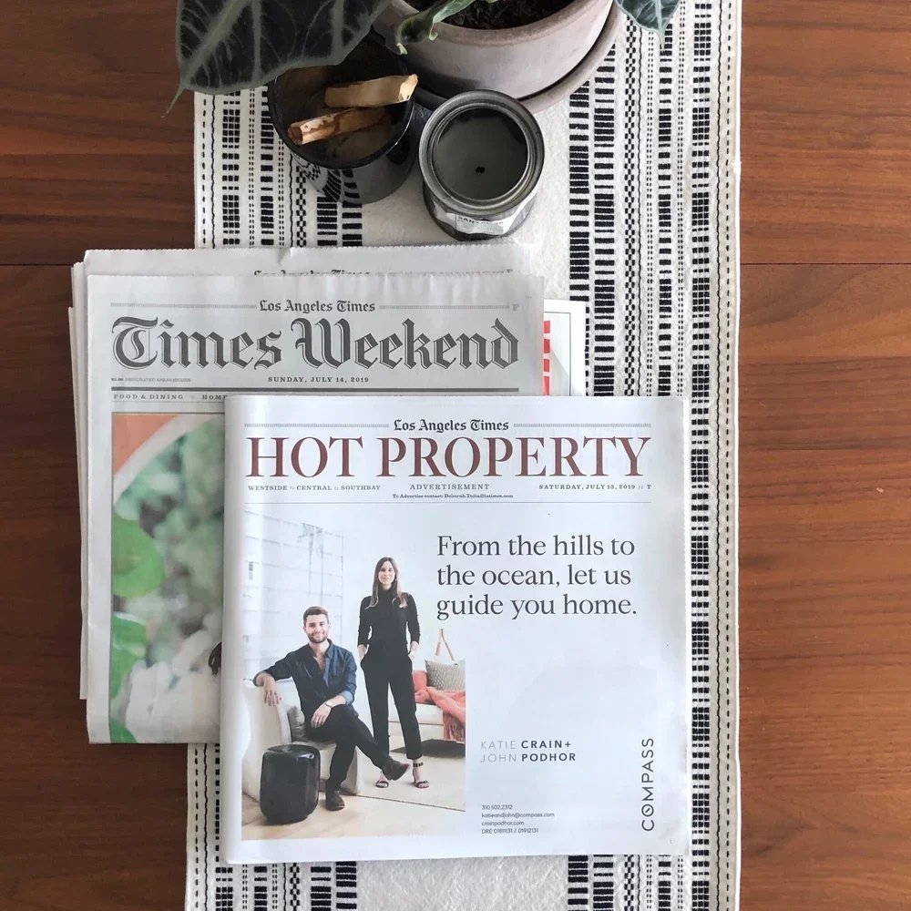 A newspaper called Los Angeles Times, specifically the section 'Hot Property,' is placed on top of a copy of the Los Angeles Times Weekend. The table has a decorative table runner underneath the newspapers. There are two candles, one black and one clear, and a potted plant on the table.