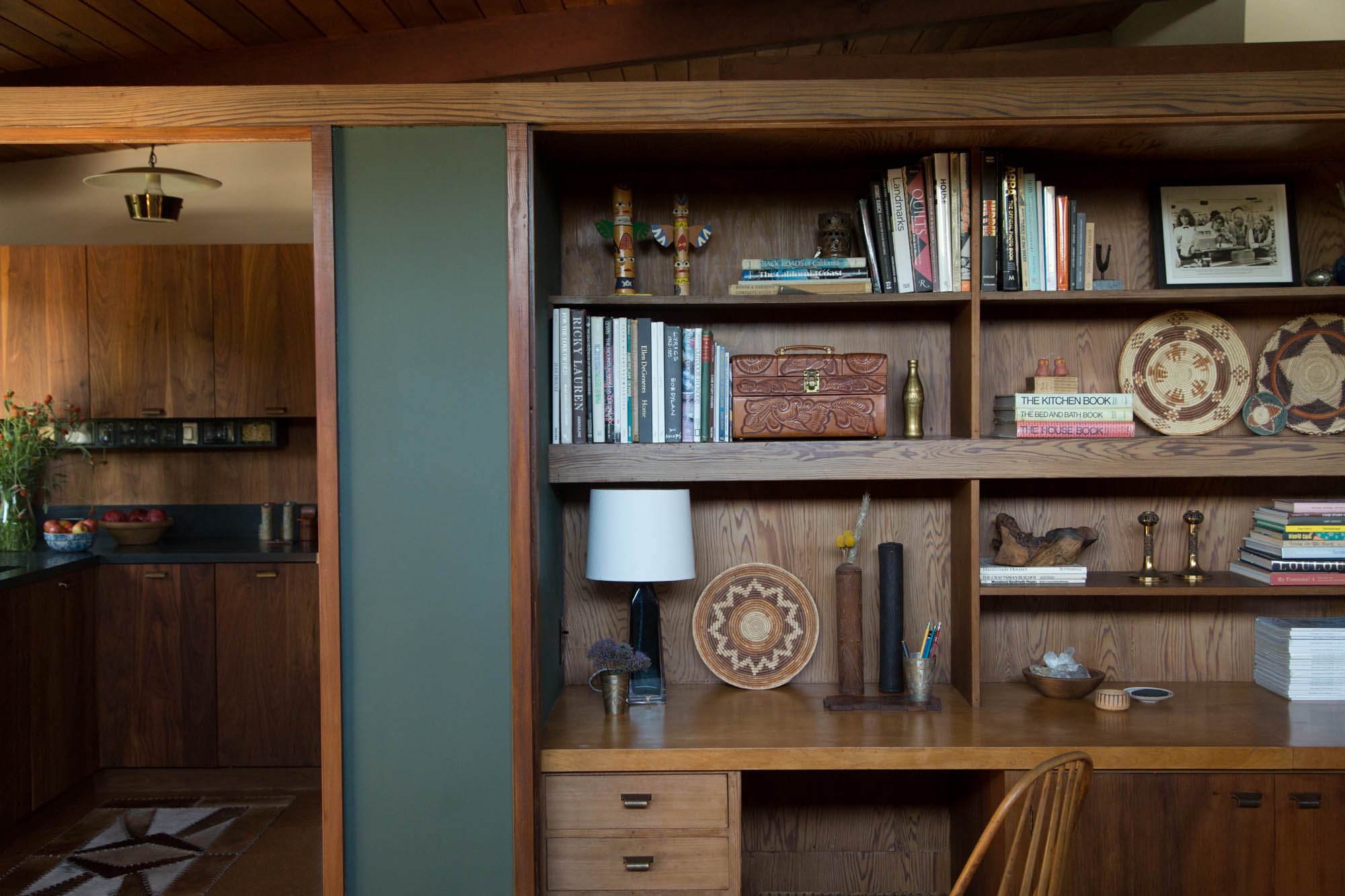 Living room with wooden shelves, books, decorative plates, and objects, with part of a kitchen visible in the background.
