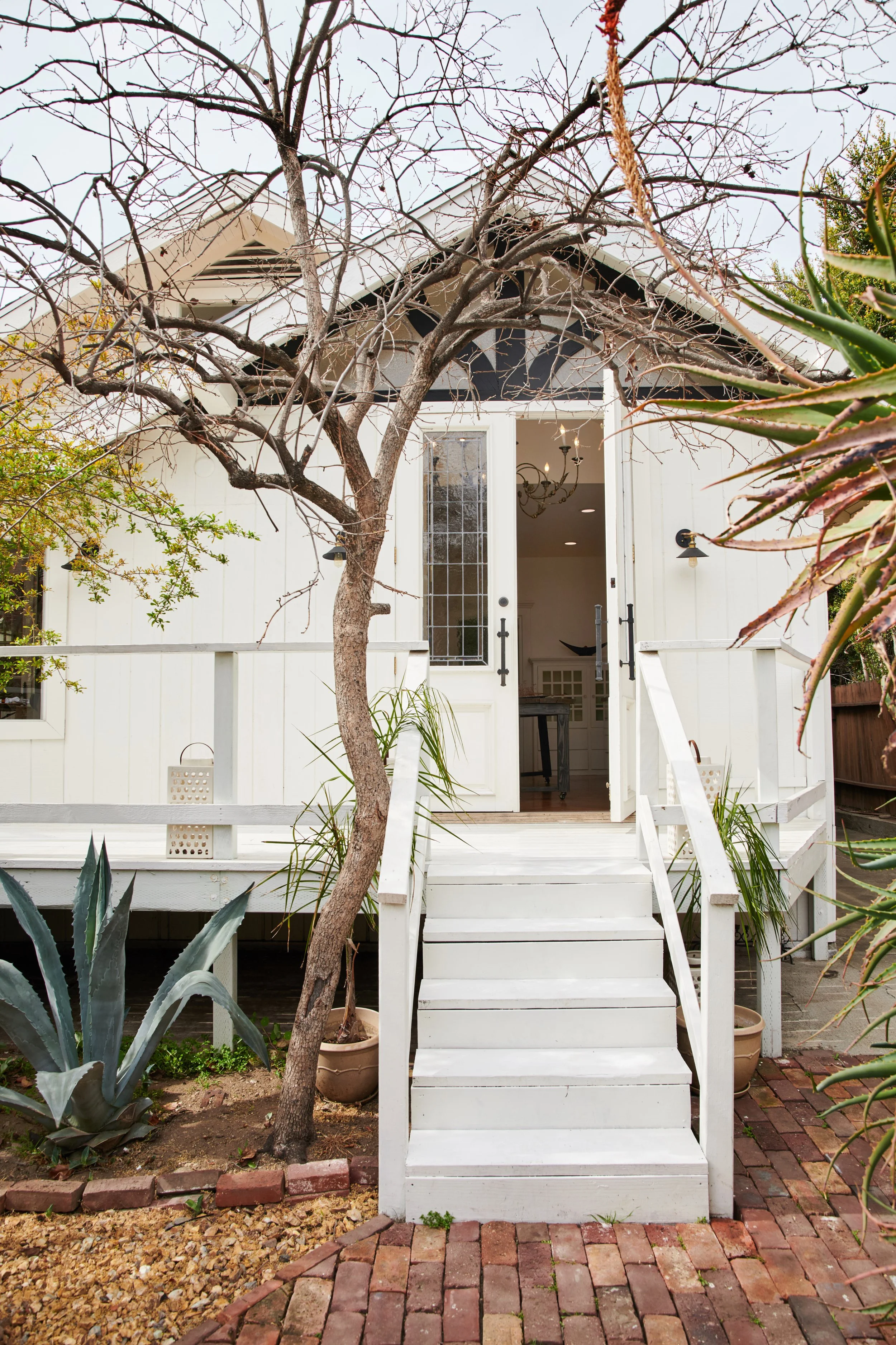 White house with open front door, staircase, and plants, including a leafless tree and aloe vera, on a brick pathway.