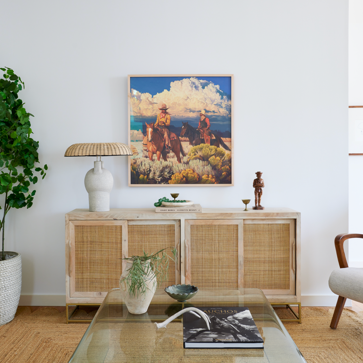Living room with a wooden sideboard decorated with a lamp, books, and small sculptures, a glass coffee table with a plant, bowl, and magazine, a large leafy plant in a basket, and a colorful landscape painting of two cowboys on horseback on the wall.