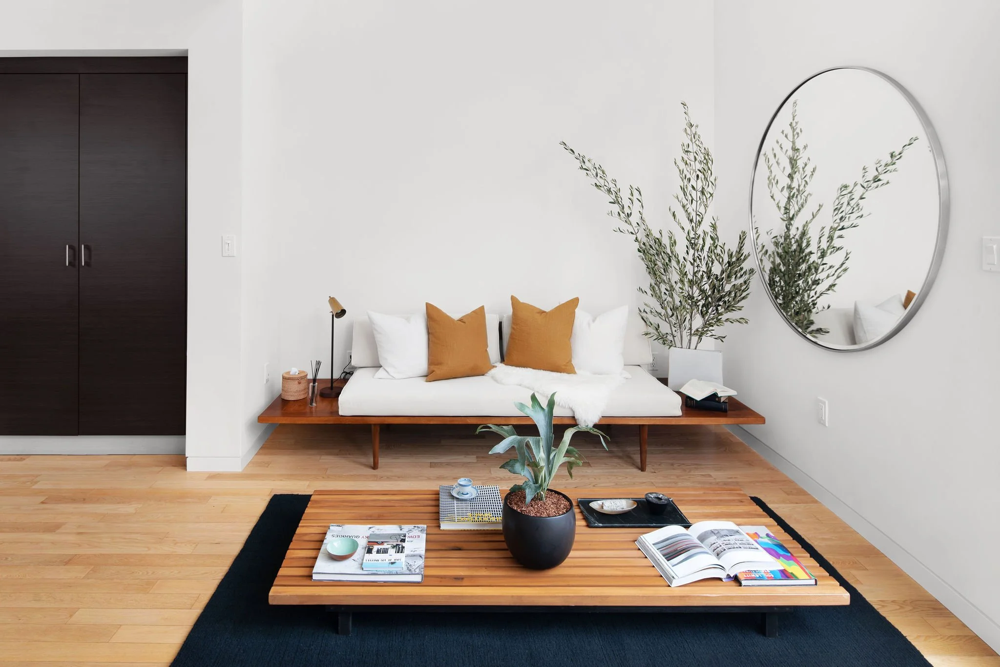 Minimalist living room with a low wooden sofa, white cushions, and orange pillows, a large potted plant on a coffee table, a round mirror on the wall, and a wooden cabinet on the left.