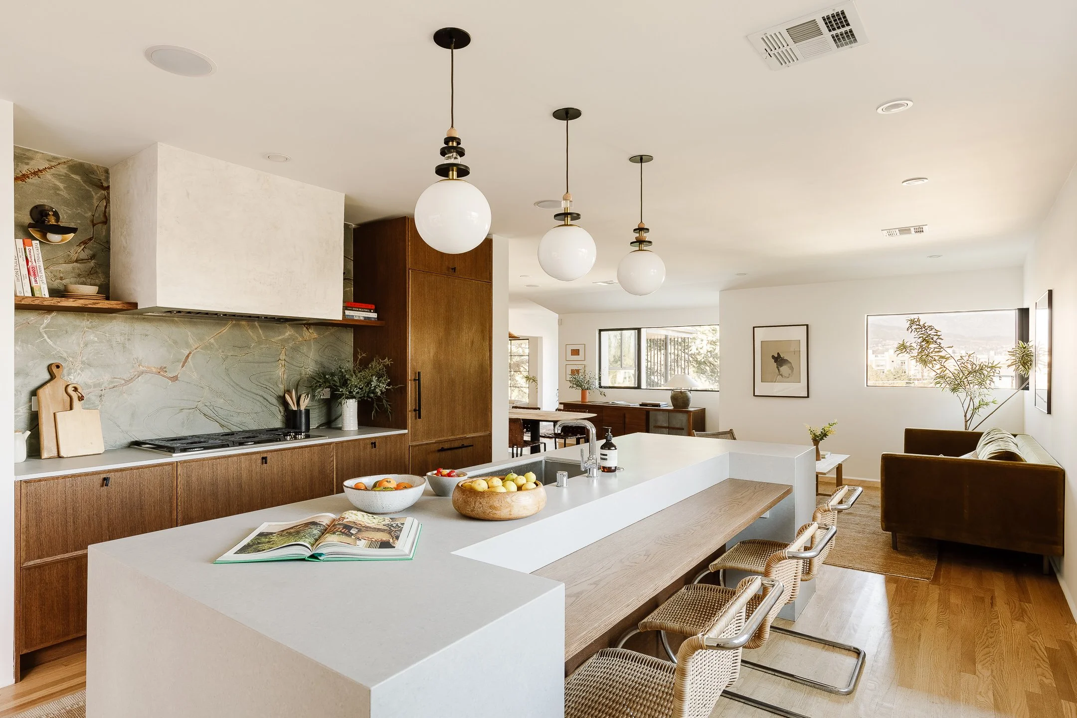 Modern open-concept kitchen and living room with white island, wooden cabinetry, three pendant lights, and natural light from large windows.