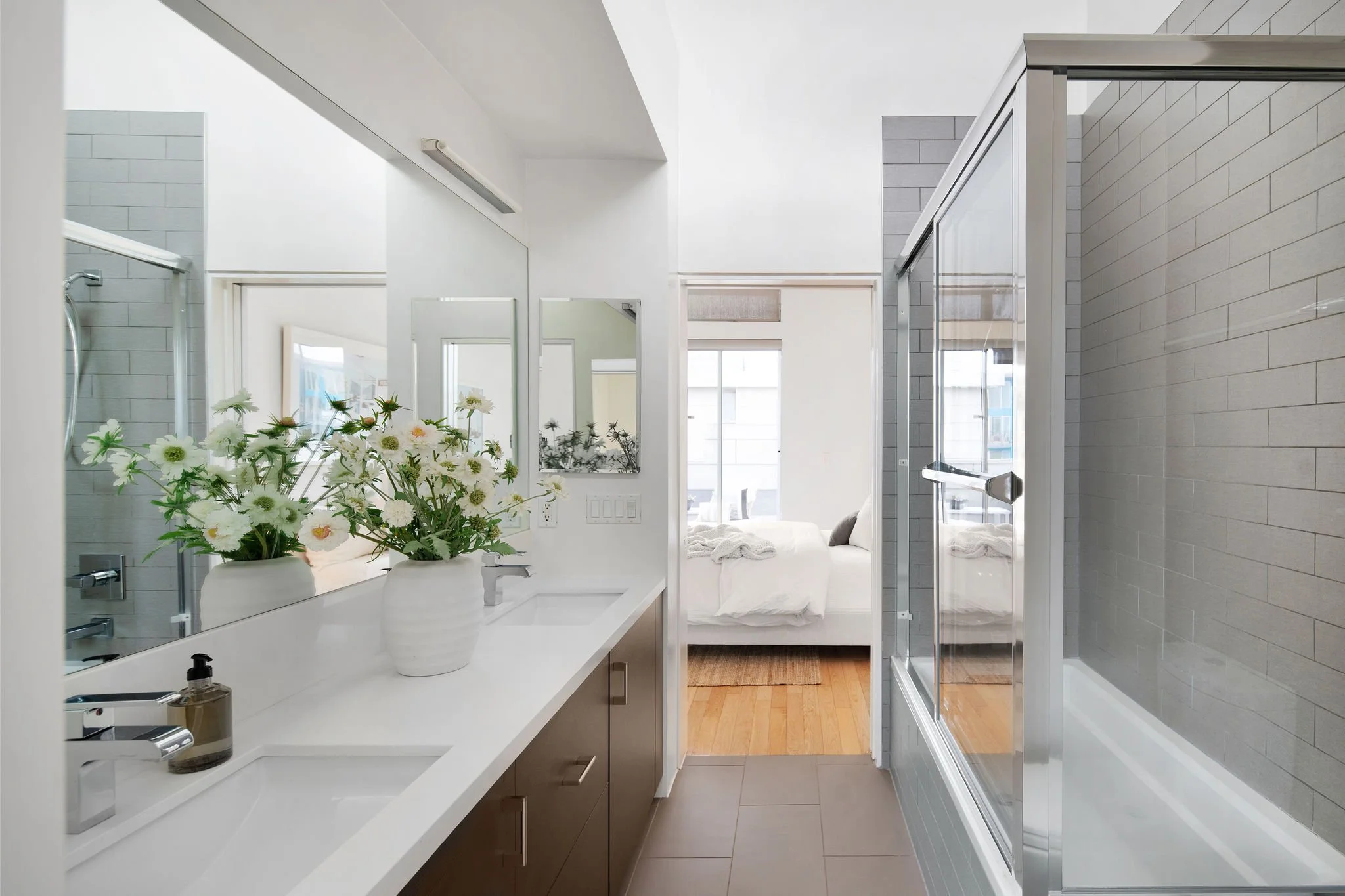 Modern bathroom with double sink vanity, large mirror, white walls, gray floor tiles, and a glass shower enclosure. Bedroom visible through doorway with bed and large window.