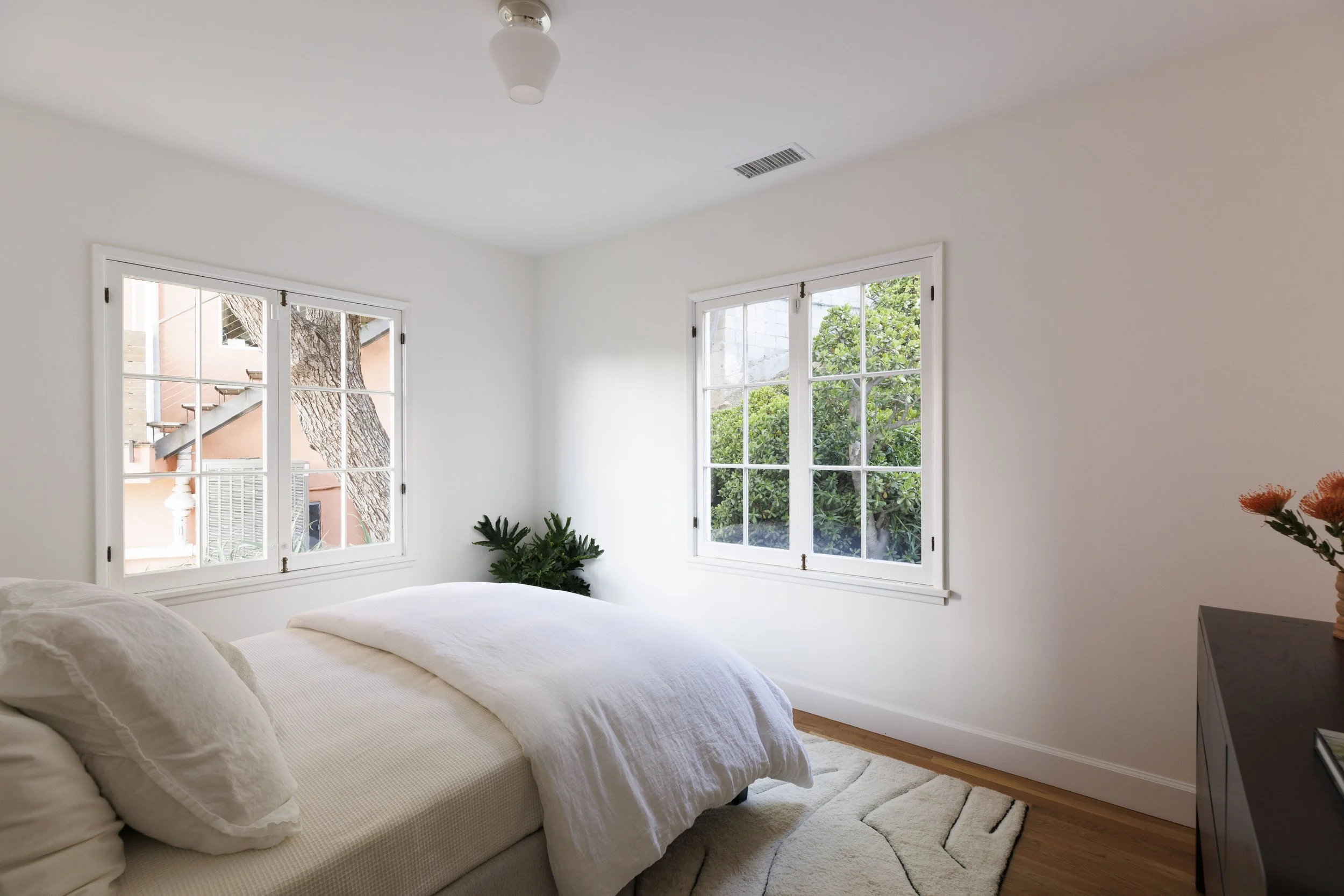 A bedroom with white walls, two large windows showing outdoor greenery, a bed with white bedding, a potted plant near the windows, a dark dresser with a vase of orange flowers, and a ceiling light fixture.