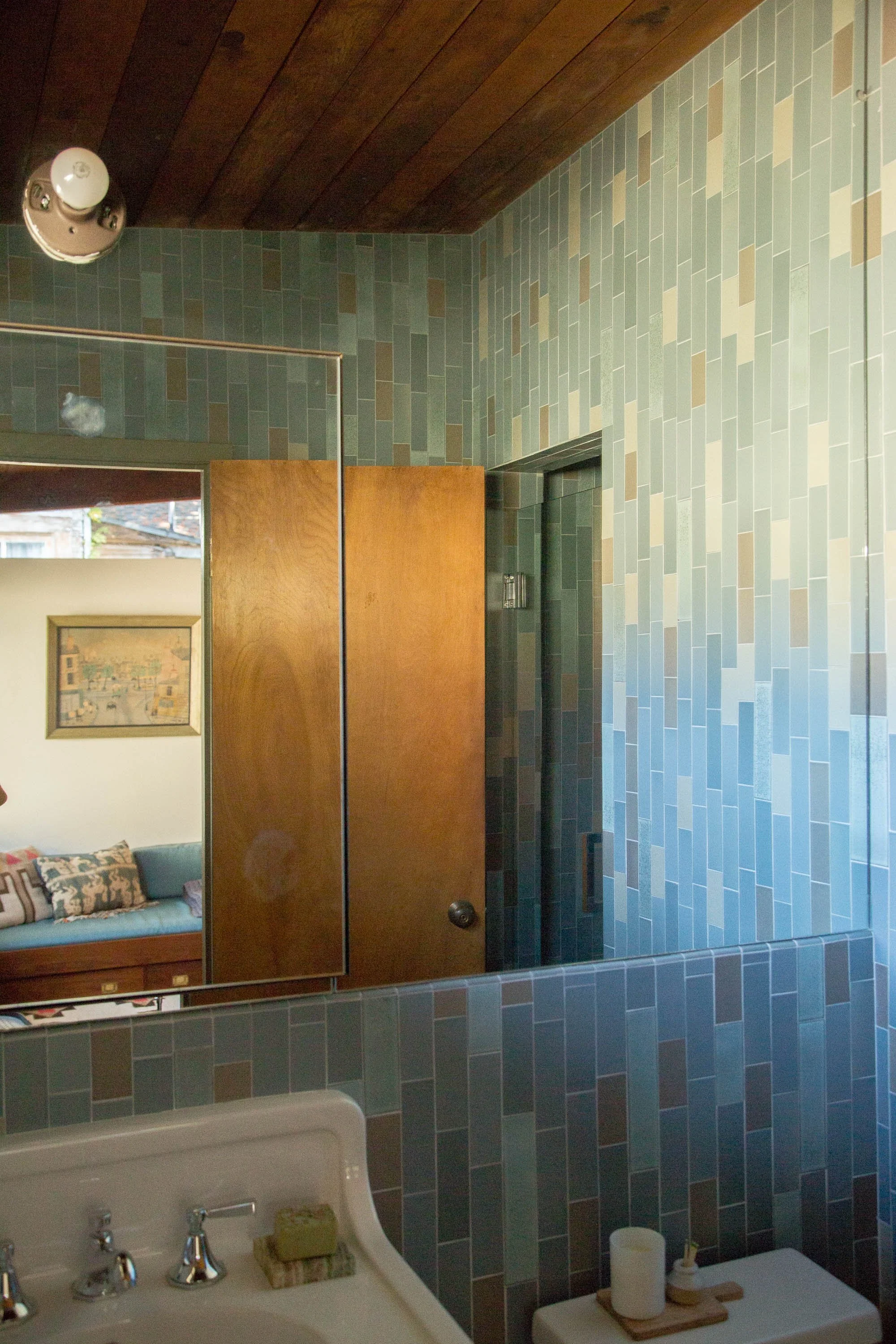 Bathroom with green and beige tile walls, wooden ceiling, mirror, small sink with soap, and a toilet with a candle and toothbrush holder.