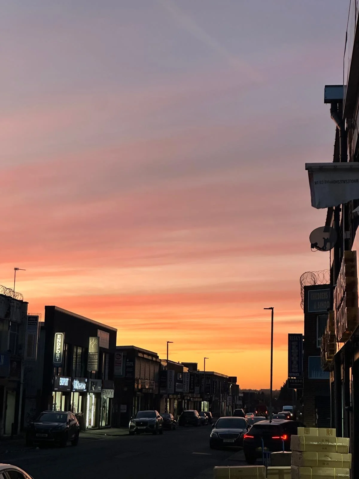 Street view at sunset with parked cars, storefronts, and a sky with colorful sunset hues.