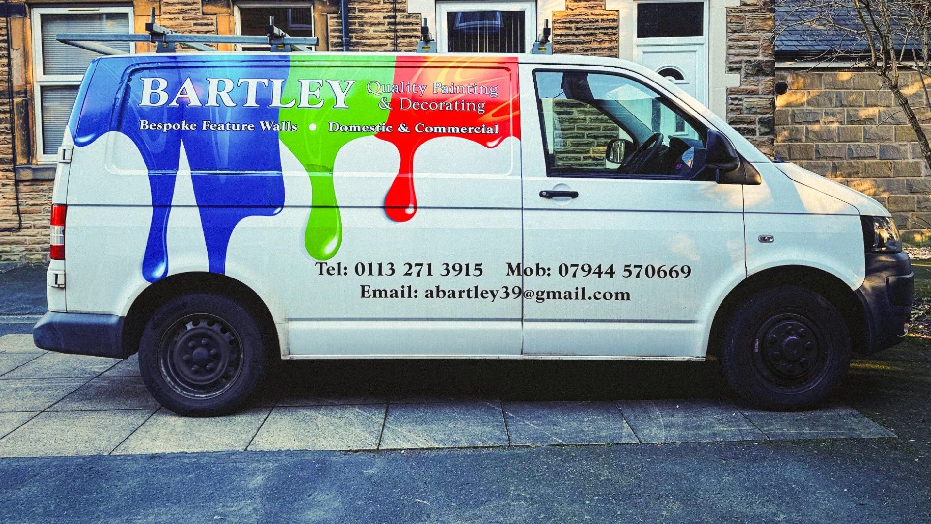 A white company van parked on a driveway in front of a stone wall house. The van has a colorful graphic design with blue, green, and red paint drips. Text on the van reads: "BARTLEY Quality Painting & Decorating" and "Bespoke Feature Walls Domestic & Commercial." Contact information includes a phone number, mobile number, and email address.