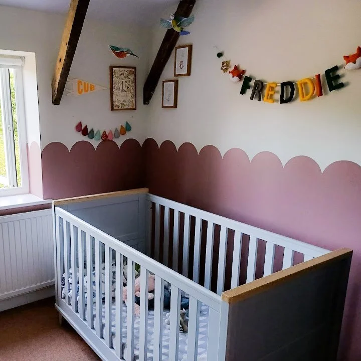 A nursery room with a white crib, a pink scalloped wall border, decorations including a banner spelling 'FREDDIE' and colorful paper hearts, and framed pictures on the wall.