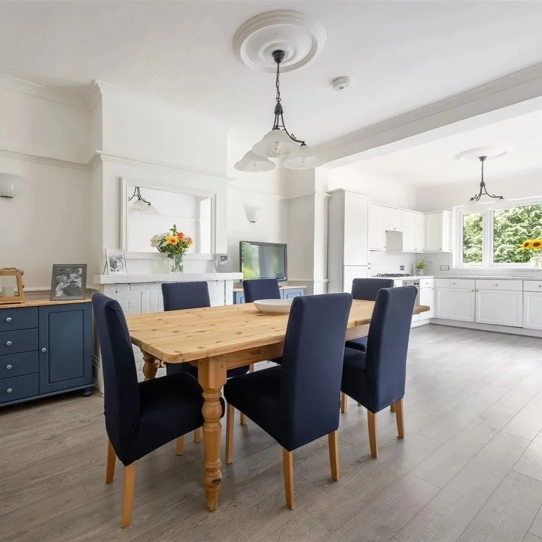 Bright dining room with a wooden table, six dark blue chairs, white walls, ceiling with two light fixtures, a blue sideboard, a mirror above the fireplace with a flower vase, a small TV, and large windows with a view of greenery.