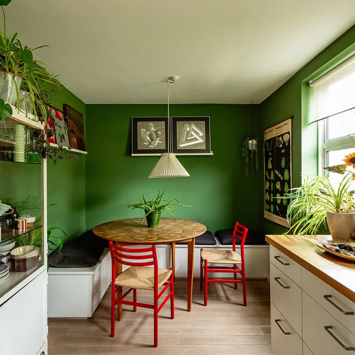 A cozy kitchen nook with a green accent wall, a round wooden table, and three chairs, two red and one beige. There are plants and artwork on the walls, a hanging light, and a window bringing in natural light.