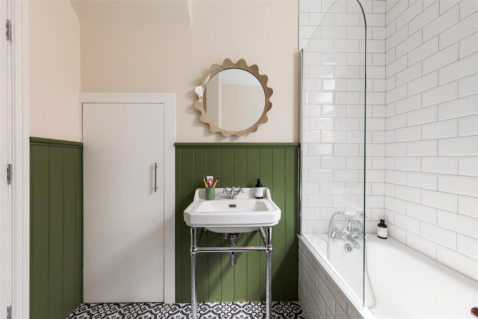 Bathroom with a white sink, green wood paneling, white tiled shower wall, and a round mirror above the sink.