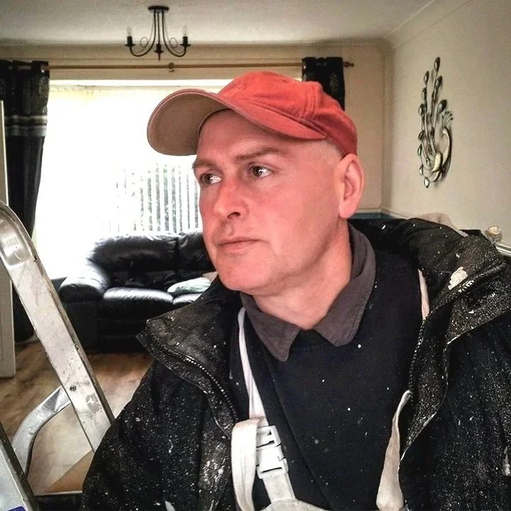 Man wearing red cap and black jacket with paint splatters, standing indoors with ladder and living room in background.