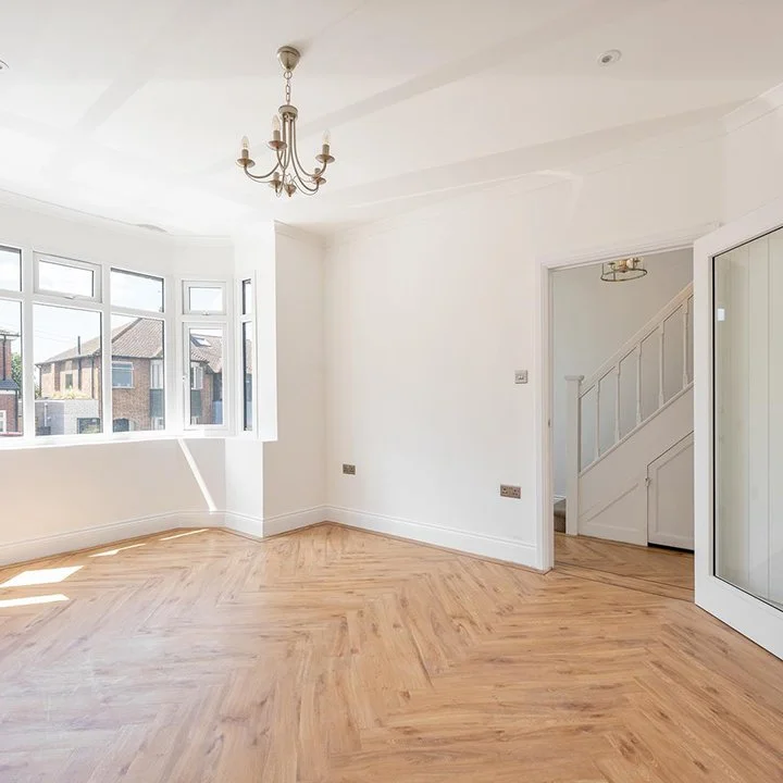 Empty room with large window, white walls, hardwood floor, chandelier, and stairs.