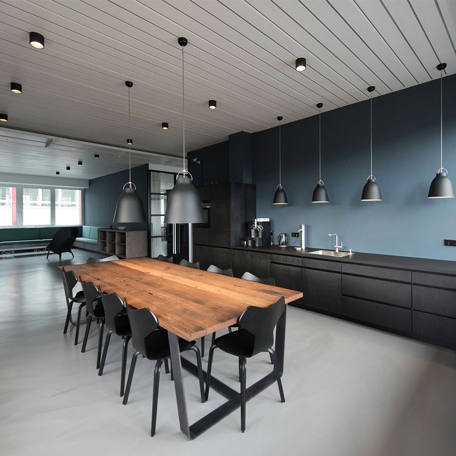 Modern kitchen and dining area with a wooden table, black chairs, and black pendant lights hanging from a white ceiling, with a seating area near windows in the background.