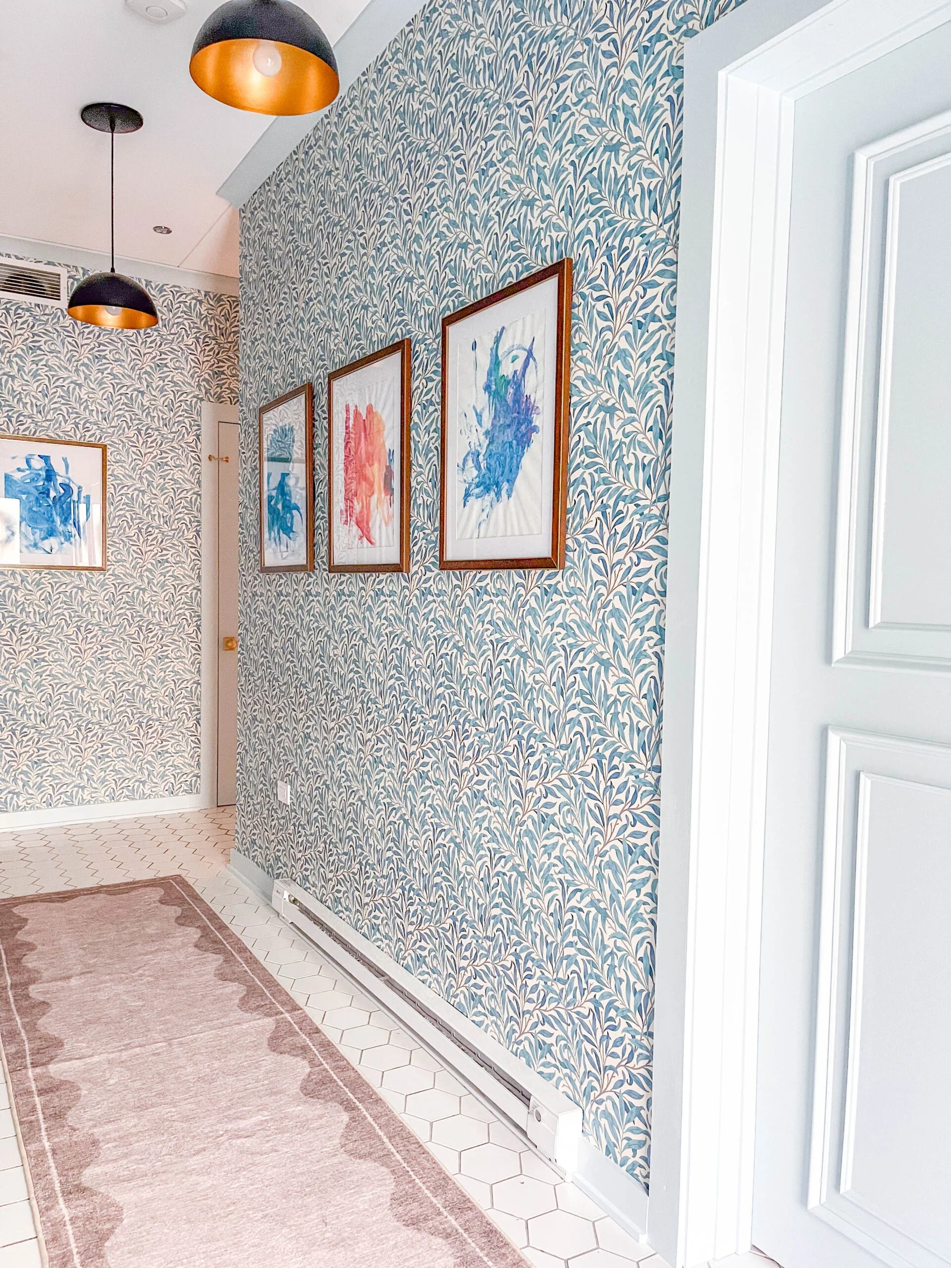 Interior hallway with wallpaper featuring a leafy pattern, framed colorful abstract artwork on the wall, a beige rug on white hexagonal tile floor, and modern black and gold pendant lights hanging from the ceiling.