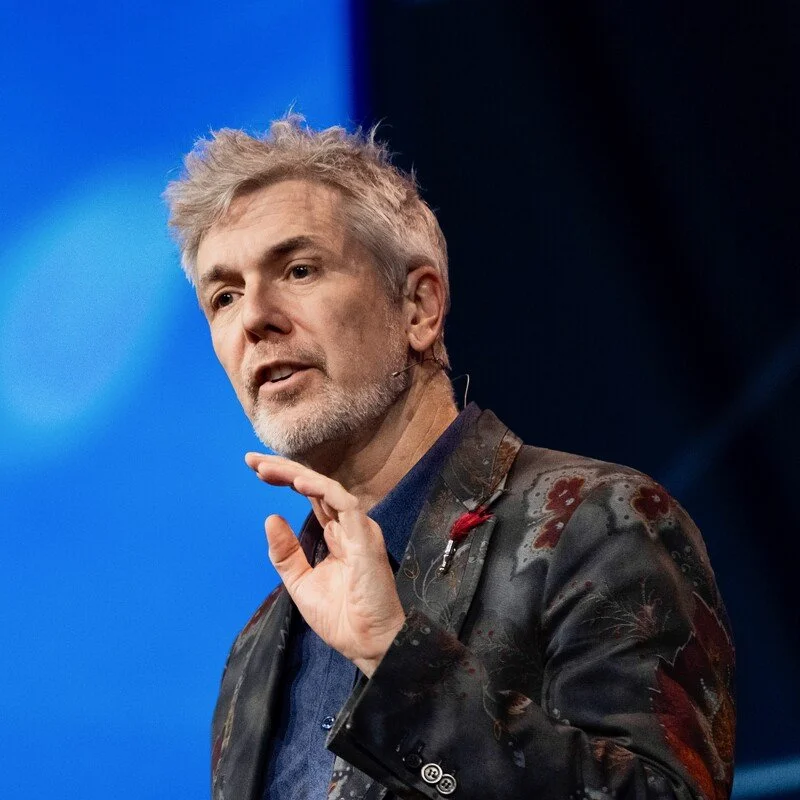 A man with gray hair and a beard, wearing a dark patterned jacket and speaking on a stage with a blue background.
