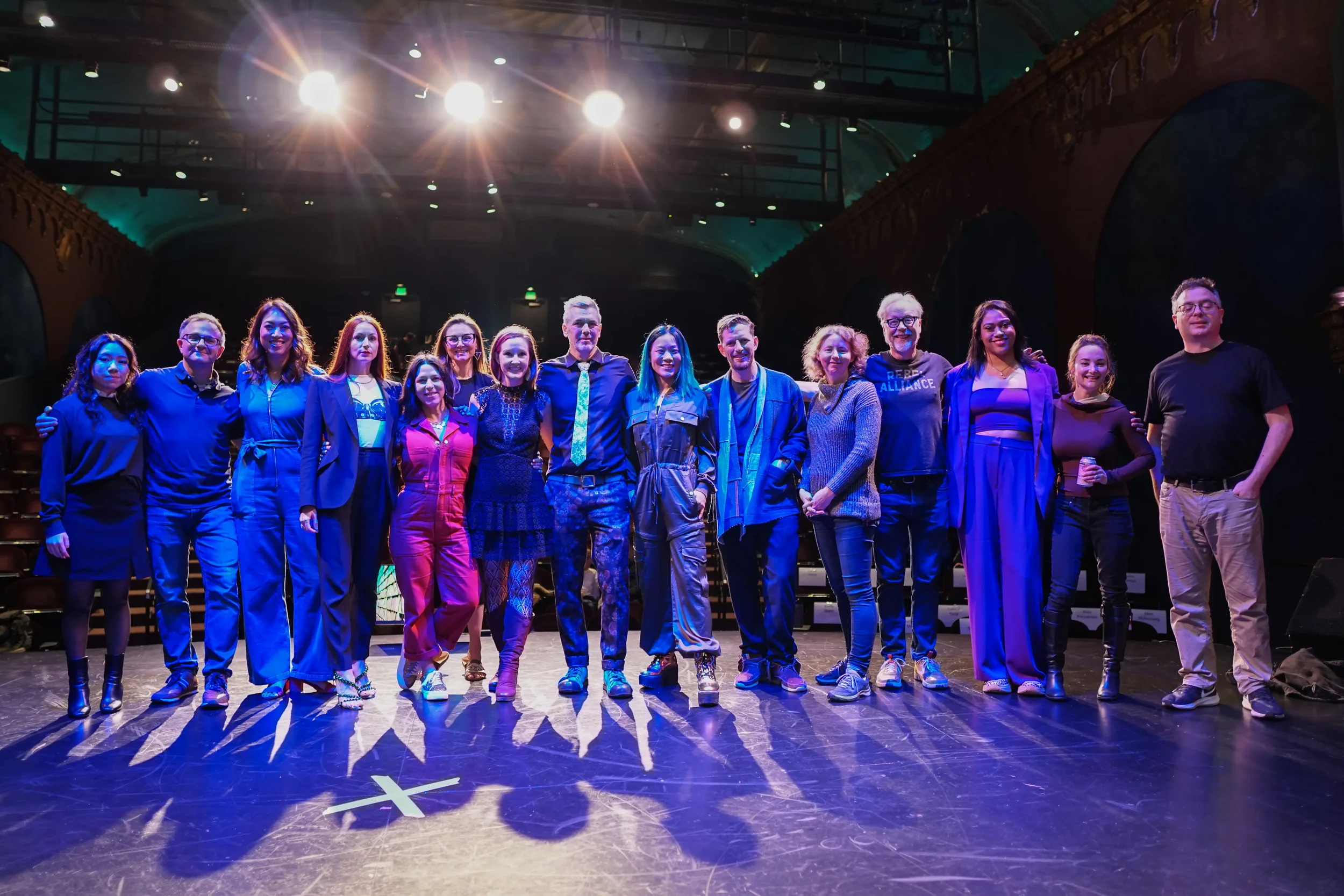 Group of 14 people standing on a stage in a theater, illuminated by stage lights, with a dark background and some upper balcony seating visible.