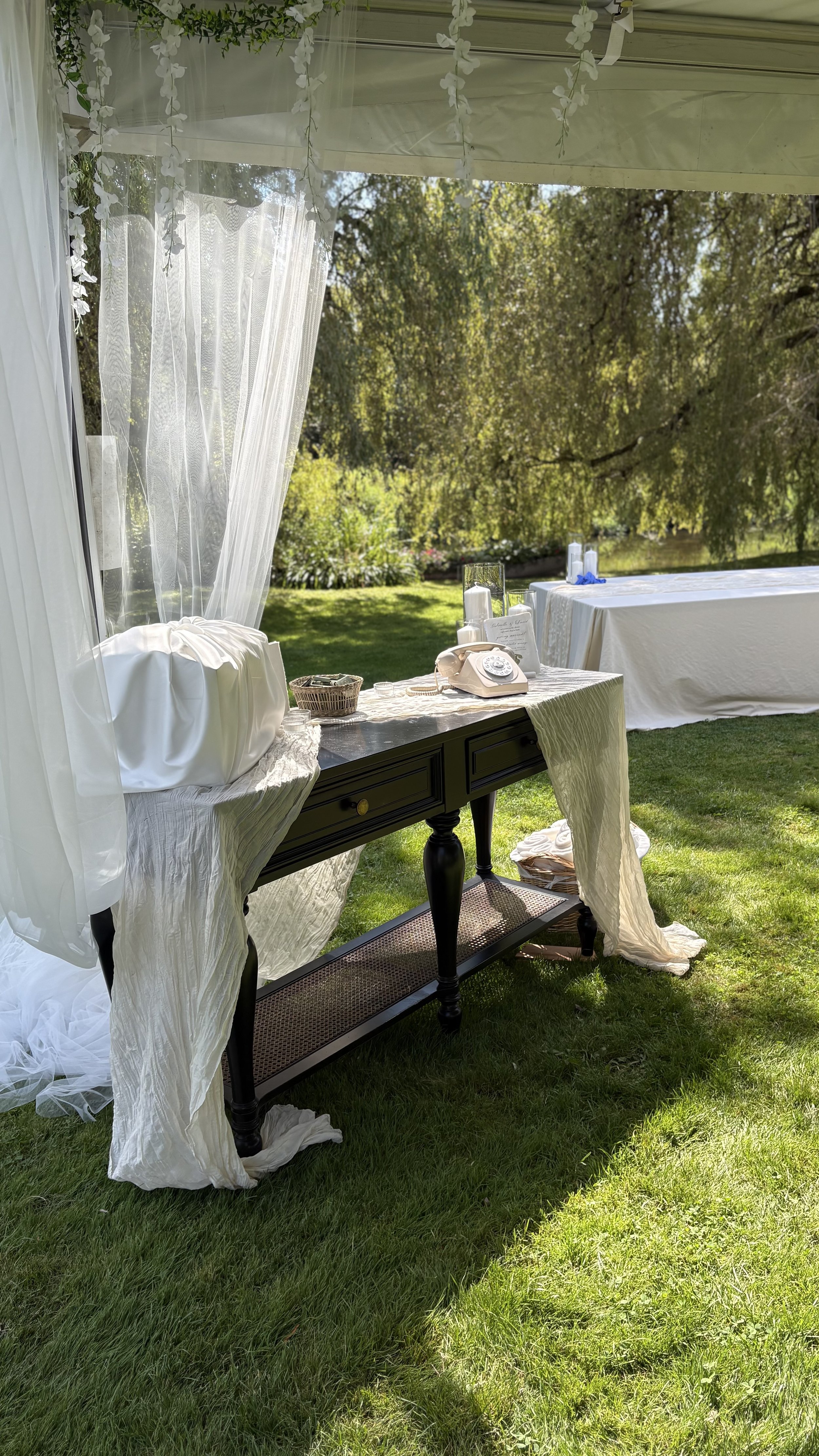 Outdoor reception table with vintage telephone, candles, and wedding decor, set in a garden under a white canopy with sheer curtains and floral decorations.