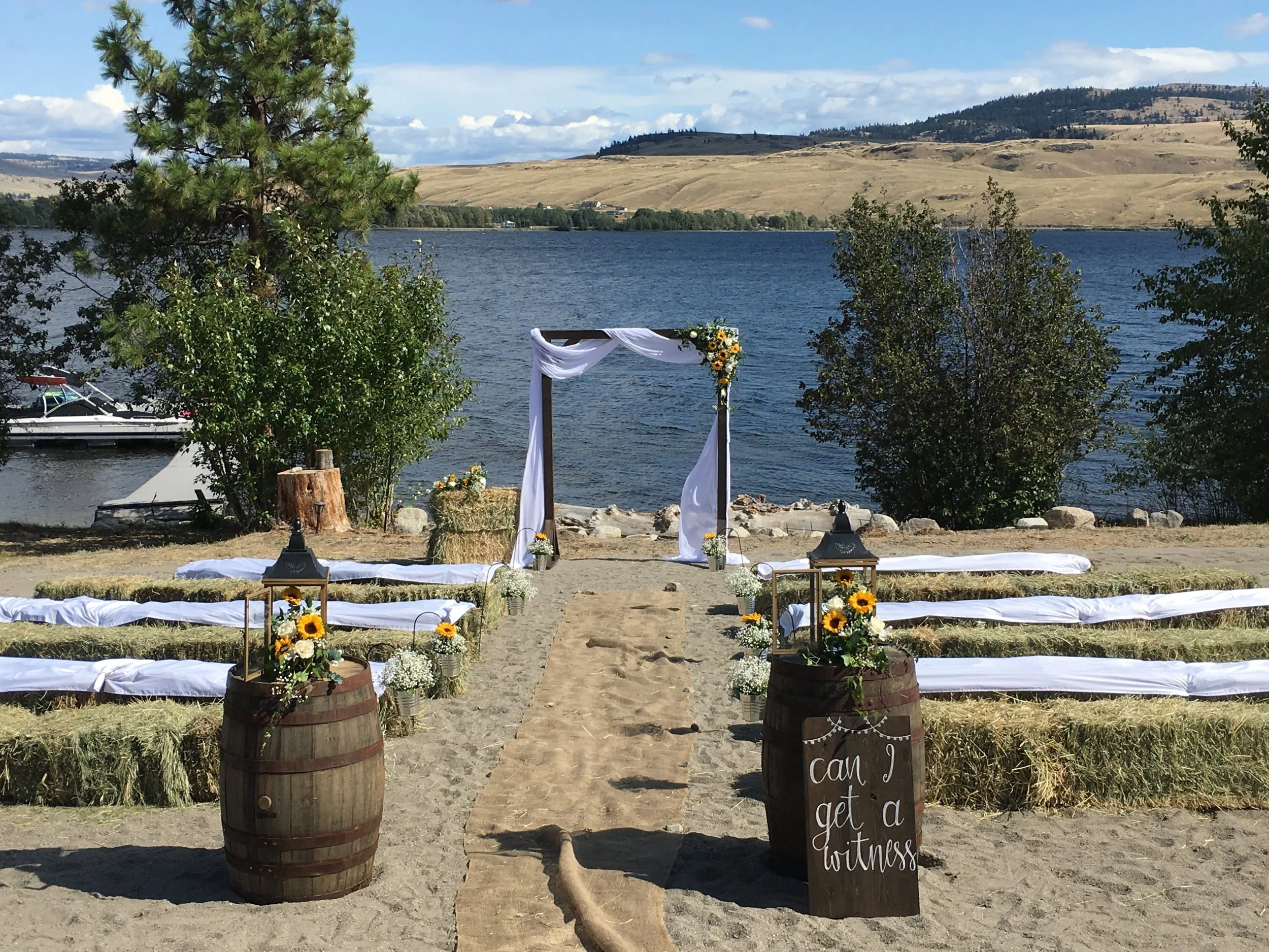 Outdoor wedding setup by a lake with an arch decorated with white fabric and flowers, wooden barrels with sunflowers, and a sign that reads 'Can I get a witness'.