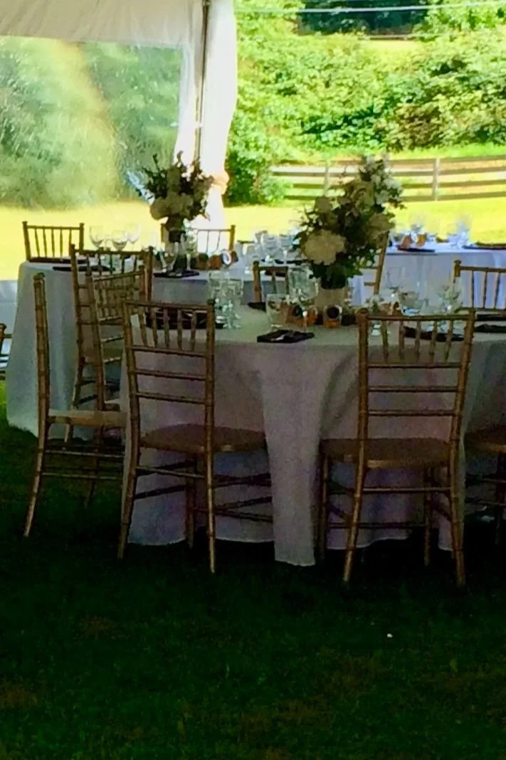 Round banquet table with white tablecloth and gold chiavari chairs, decorated with floral centerpieces and table settings, set outdoors on a grassy area with a tent and green trees in the background.