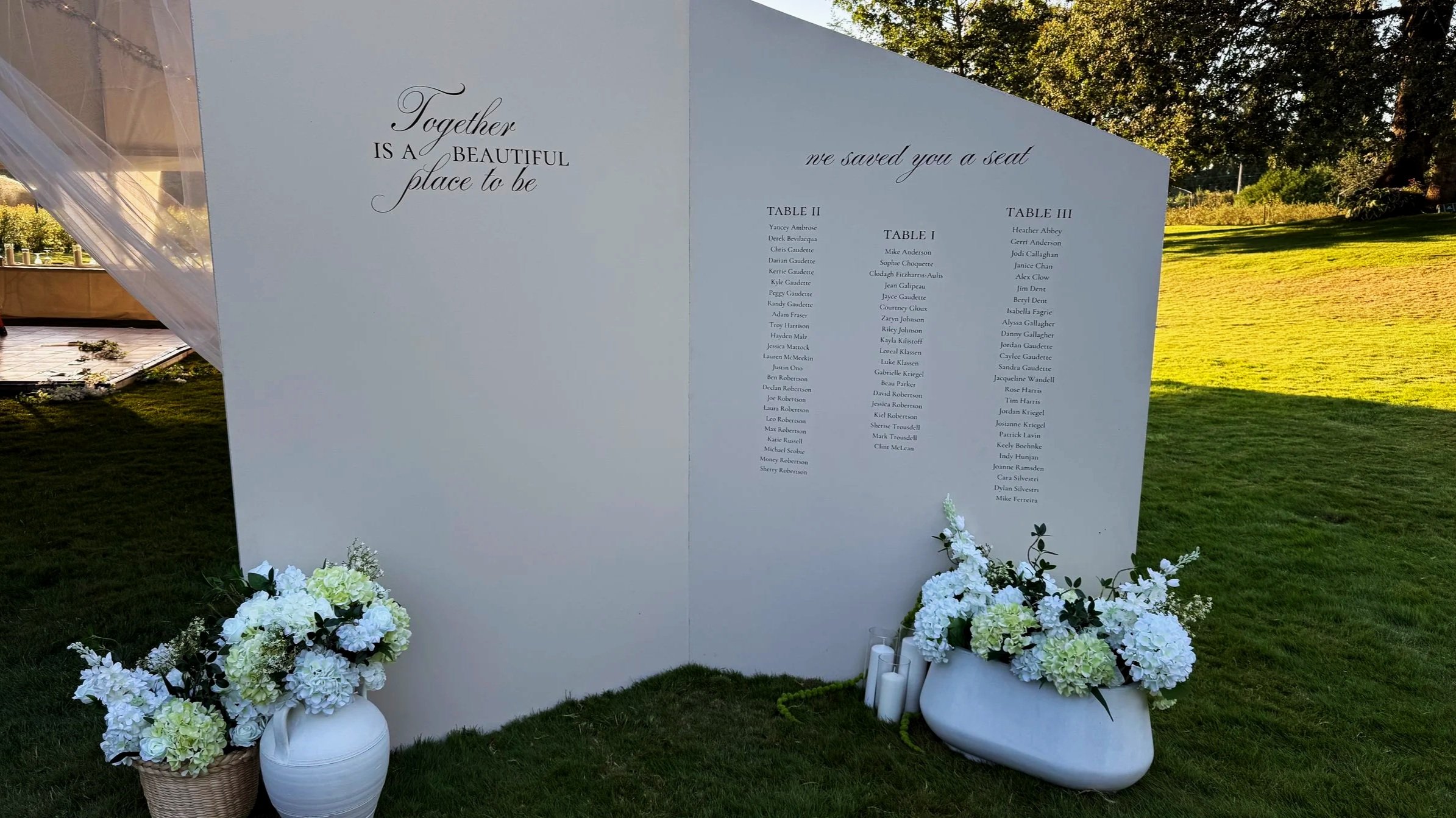 Wedding seating chart board with white floral arrangements at the base, set outdoors on a grassy area.