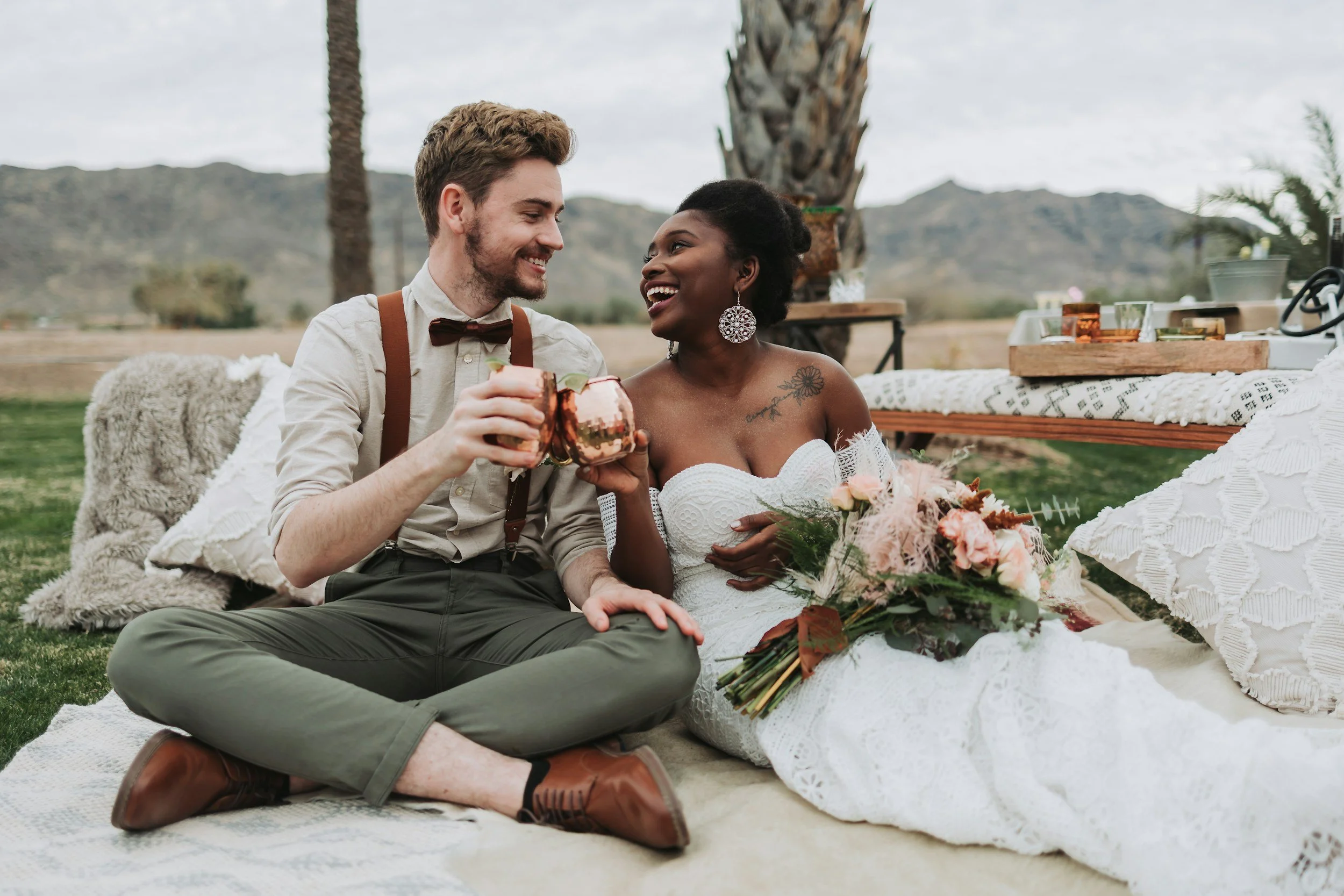 A wedding couple sitting on the grass outdoors with mountains and palm trees in the background. The groom, a man with light skin, brown hair, and a beard, is wearing a light shirt with suspenders and a bow tie. The bride, a woman with dark skin and black hair styled up, is dressed in an off-the-shoulder white lace wedding dress and holding a bouquet of flowers. They are smiling and toasting with metallic cups, enjoying a celebratory moment together.